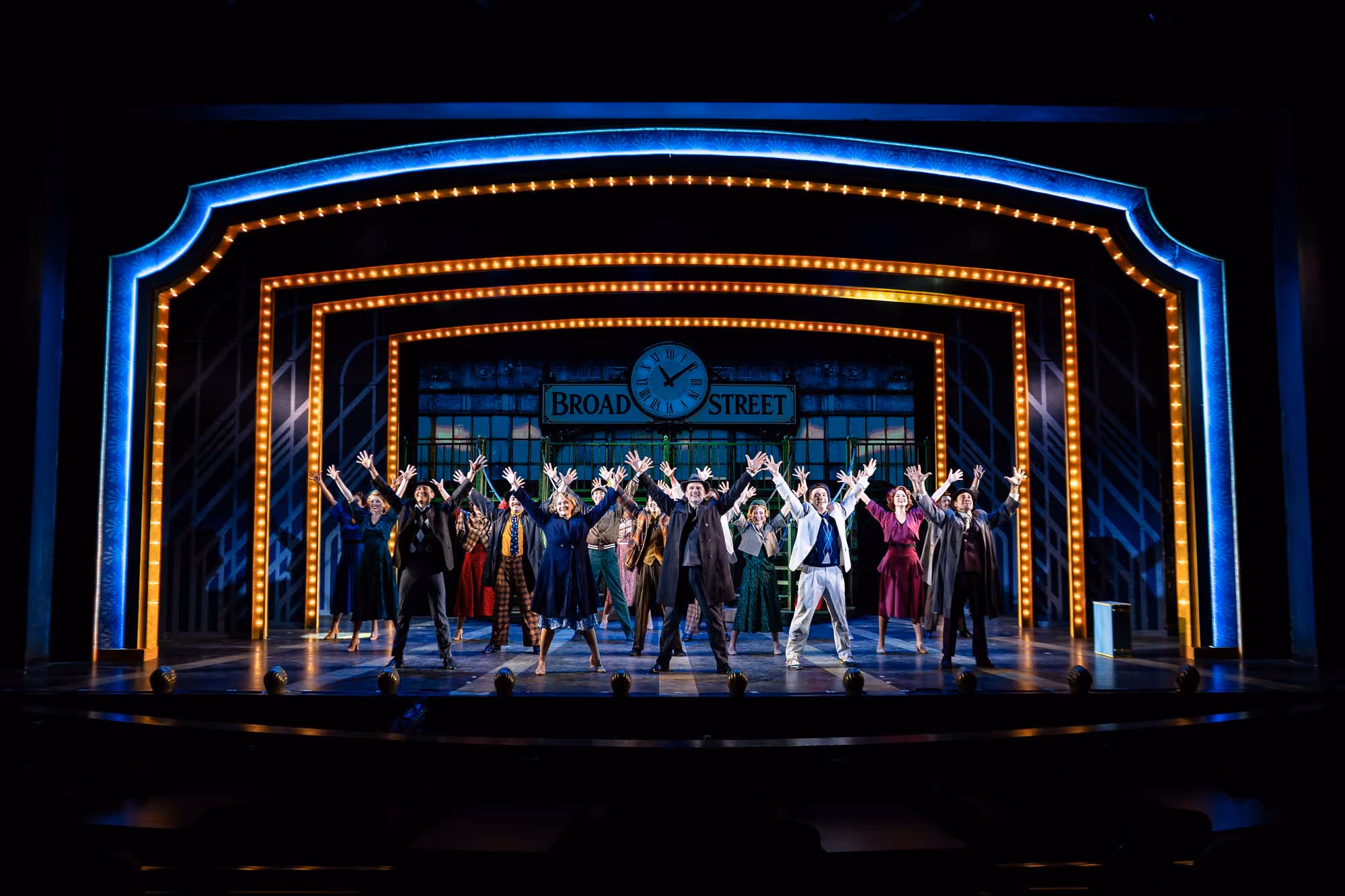 Stage performance with a group of actors raising their hands, framed by illuminated arches and a sign reading Broad Street.