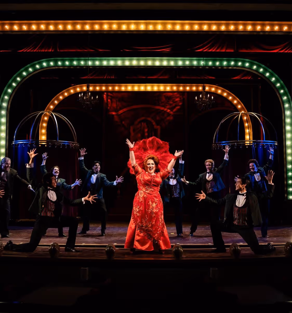 Theatrical performance with a woman in a vibrant red gown center stage surrounded by men in tuxedos striking dramatic poses under colorful lit arches.