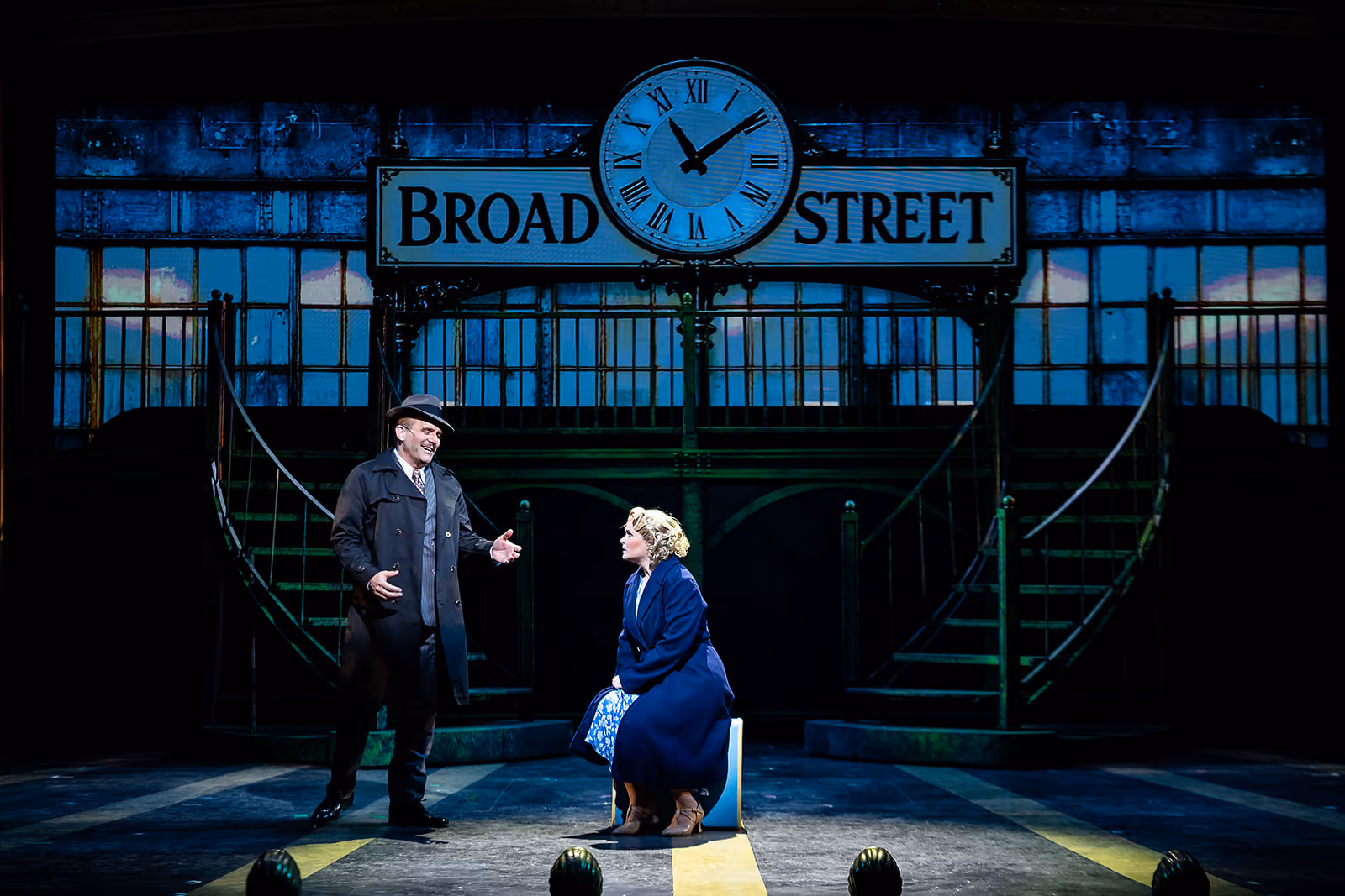 A man in a dark coat and hat stands speaking to a seated woman in a blue coat on a stage set depicting a Broad Street subway station with a large clock showing 2:05.
