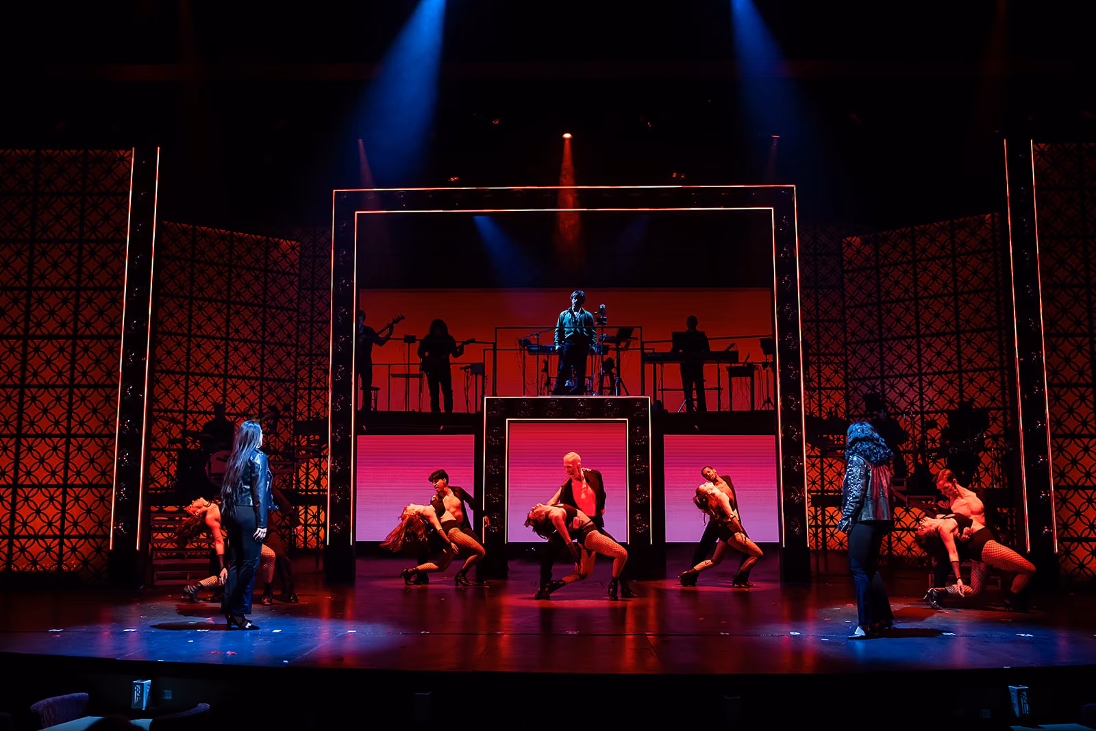 A theatrical stage performance featuring several dancers and actors under dramatic lighting with a musician elevated in the center background against a red-lit backdrop framed by geometric lighted structures.