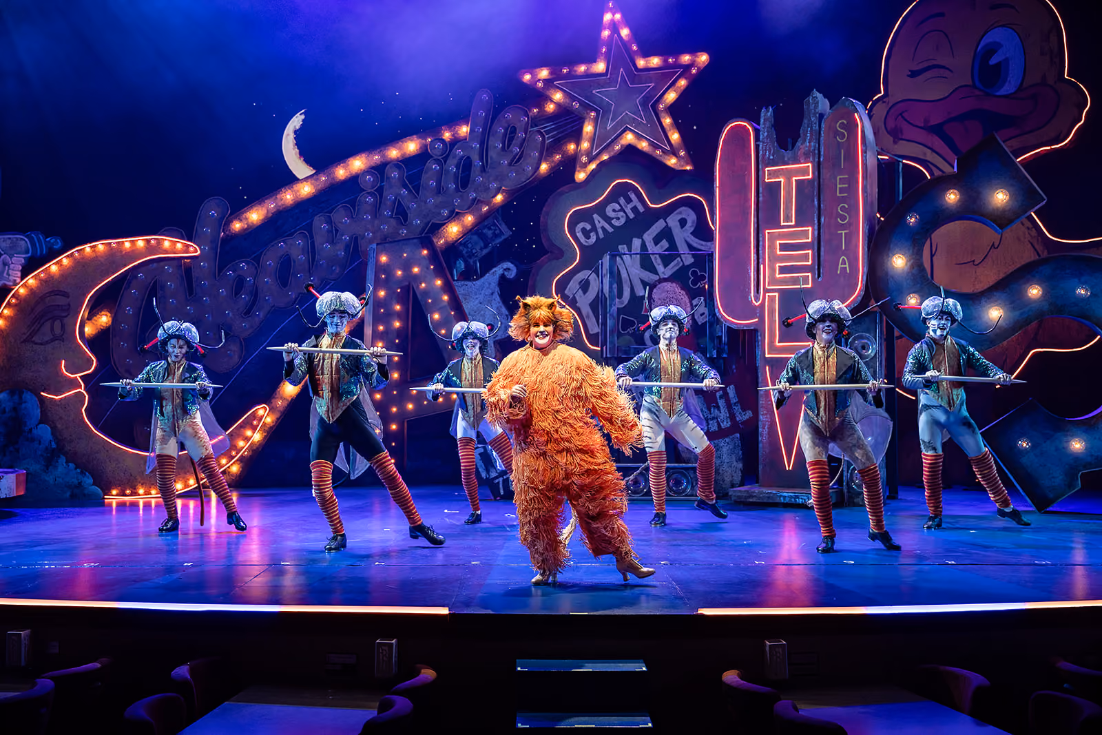A theatrical stage performance with a person in an orange furry animal costume standing center front and six performers behind them dressed in colonial-style outfits holding long sticks, colorful neon signs in the background including words like 'Cash Poker' and 'Siesta'.