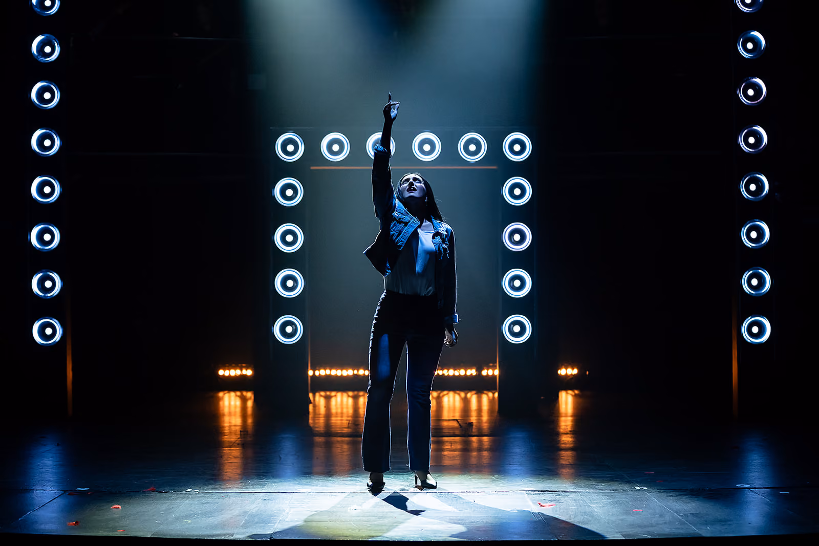 A female performer stands center stage under a spotlight, pointing upward with one hand, surrounded by circular stage lights forming a rectangle on a dark theater stage.