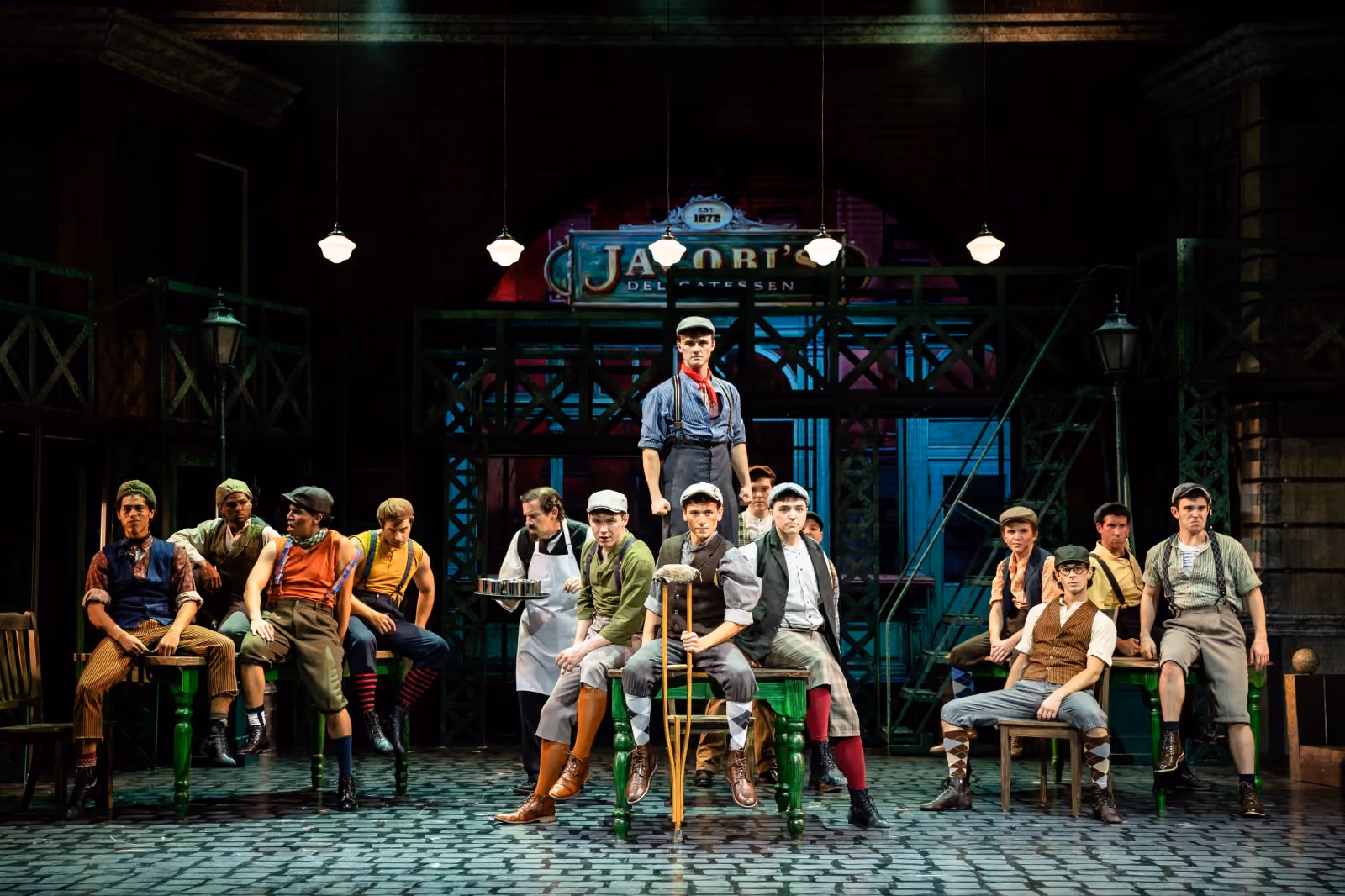 Group of young men in early 20th century newsboy costumes posing on a stage with an industrial backdrop and hanging lights.