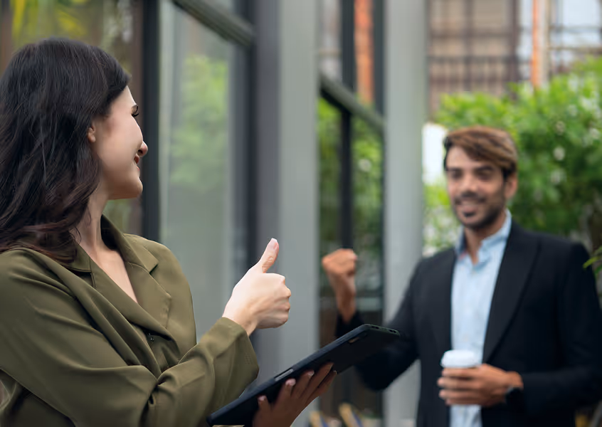 Woman showing thumbs up to a smiling man holding a coffee cup outside an office building.