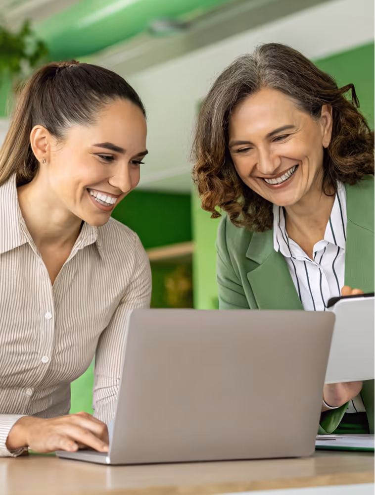 Two women smiling and looking at a laptop screen together in a bright office setting.