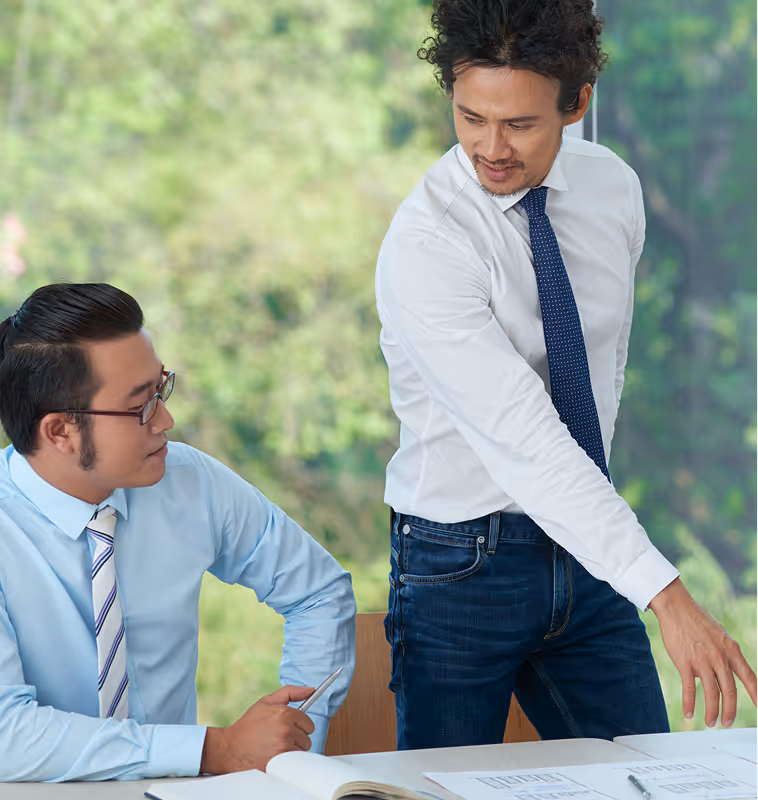 Two men in business attire discussing documents on a table, with one standing and pointing while the other sits holding a pen.