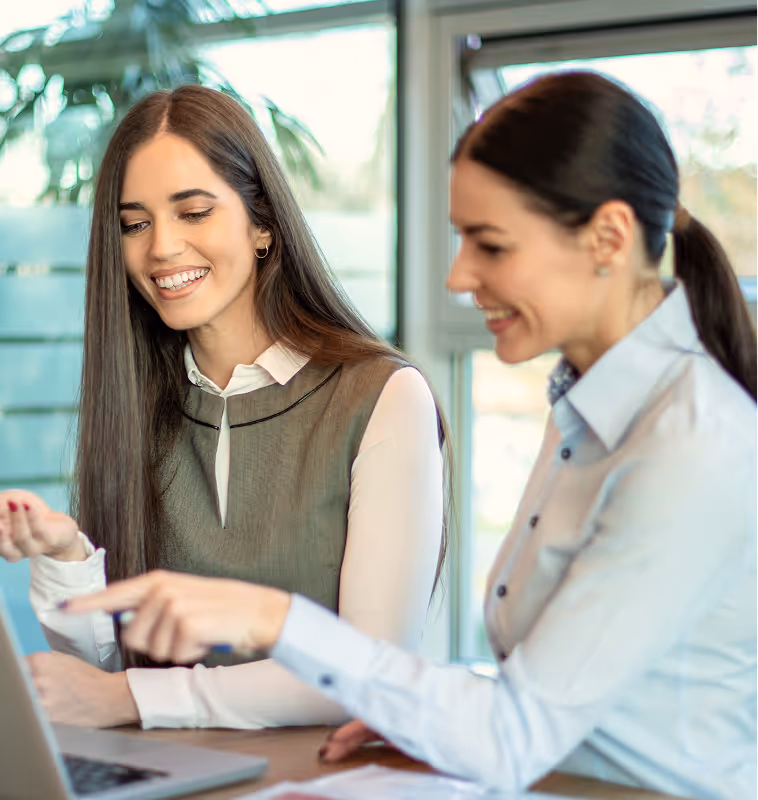 Two women smiling and looking at a laptop screen, one pointing at the screen during a discussion.
