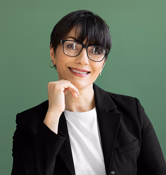 Smiling woman with short dark hair, wearing glasses and a black blazer over a white top, posing with her hand near her chin against a green background.