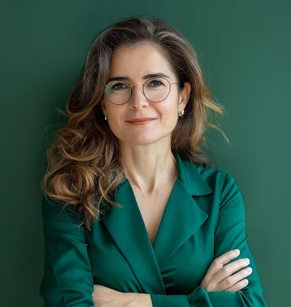 Confident woman with long wavy hair and glasses wearing a green blazer, standing with crossed arms against a green background.
