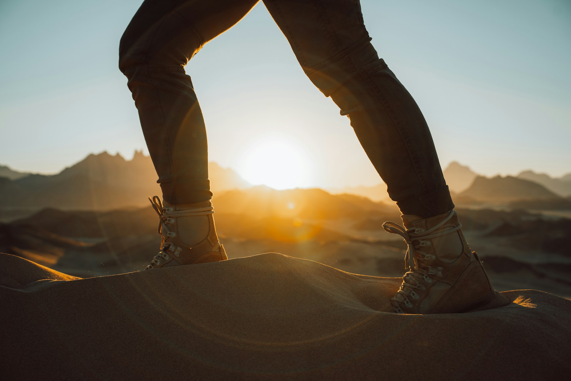 Close-up of person wearing boots standing on sand dune with mountains and setting sun in the background.