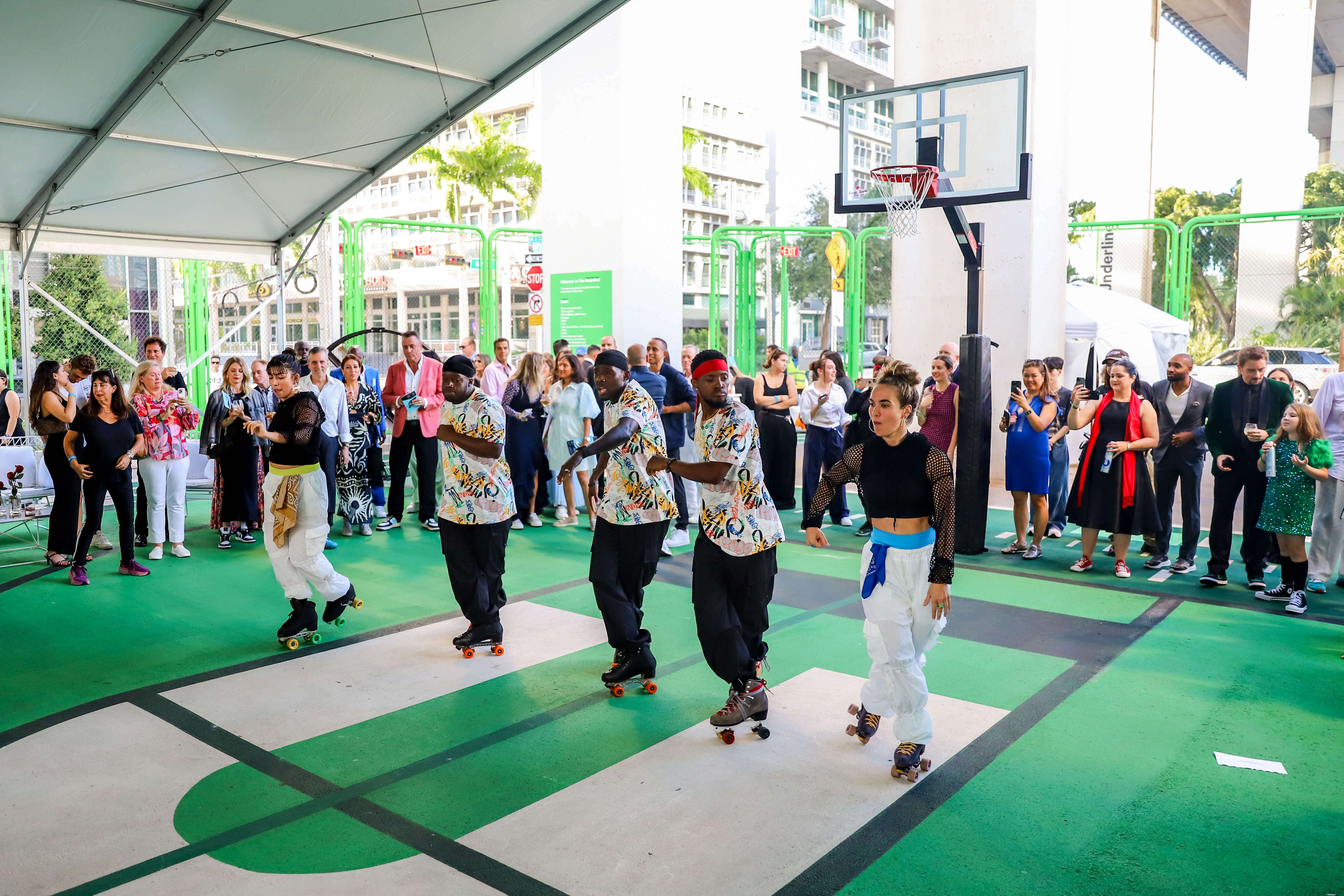 Five roller skaters performing a synchronized routine on a green outdoor basketball court with an audience watching under a large tent.