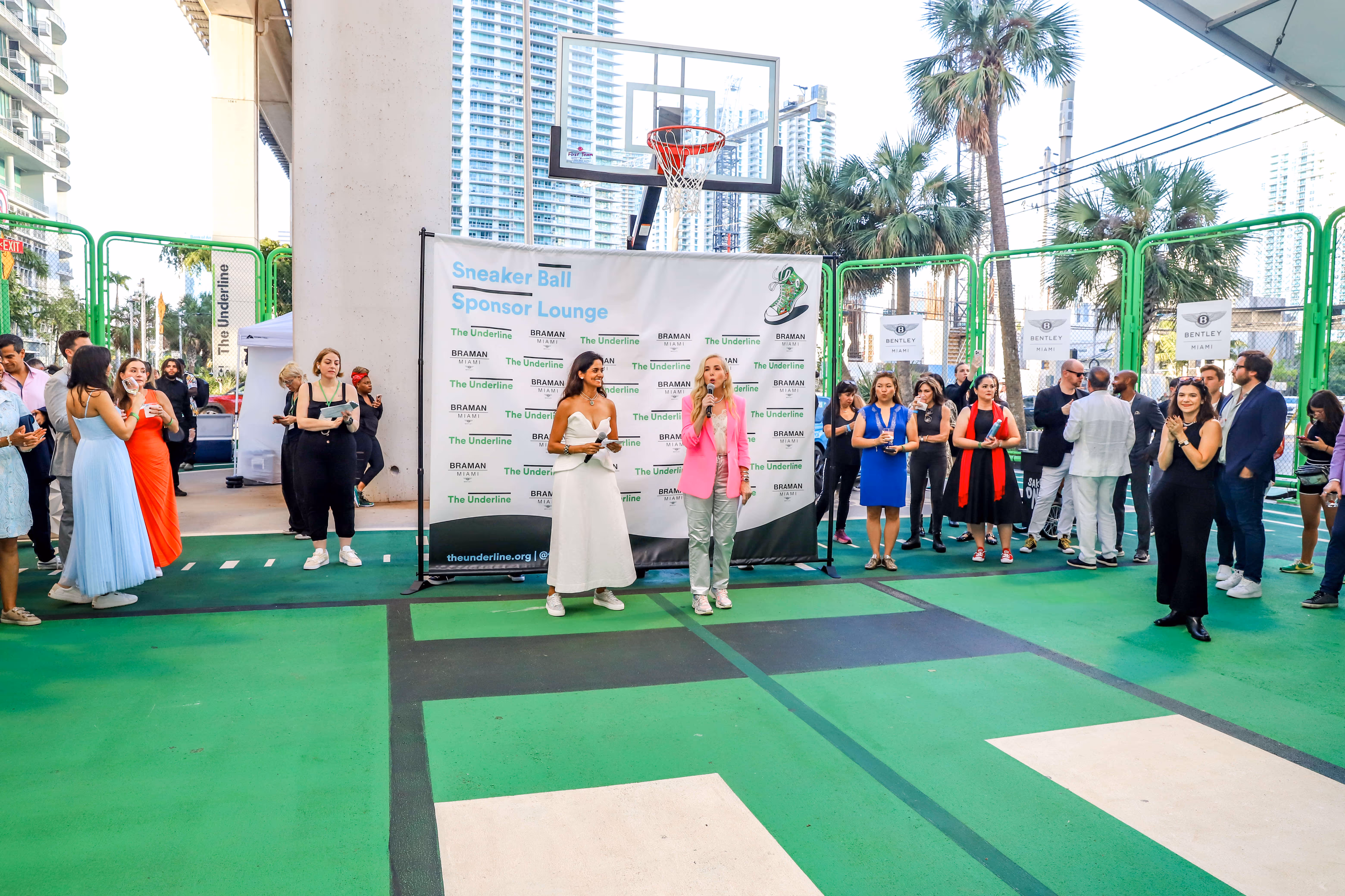 Two women speaking on a basketball court during the Sneaker Ball Sponsor Lounge event, with a crowd watching and a branded backdrop behind them.