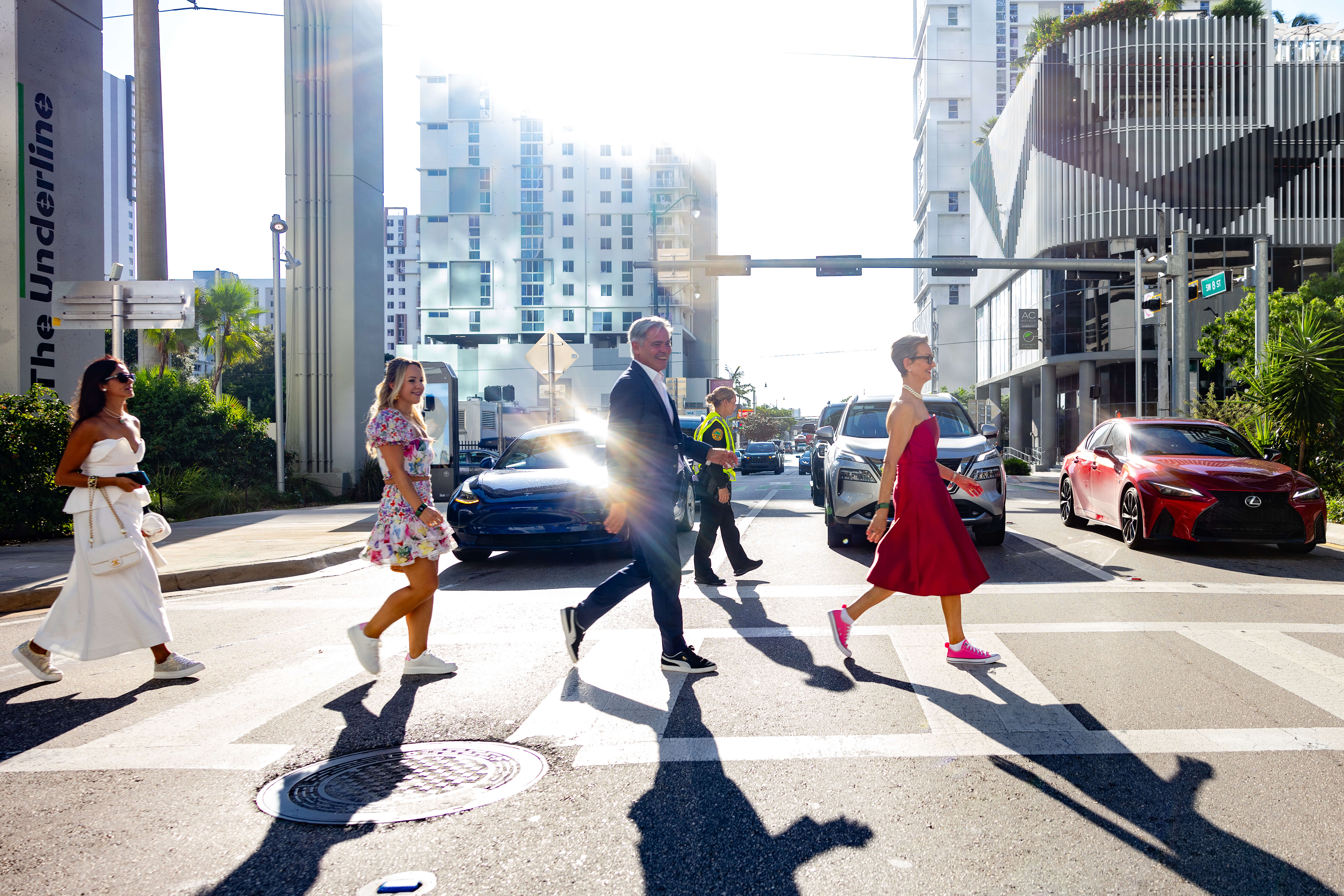 Four people walking across a city street crosswalk on a sunny day, with cars waiting and tall buildings in the background.