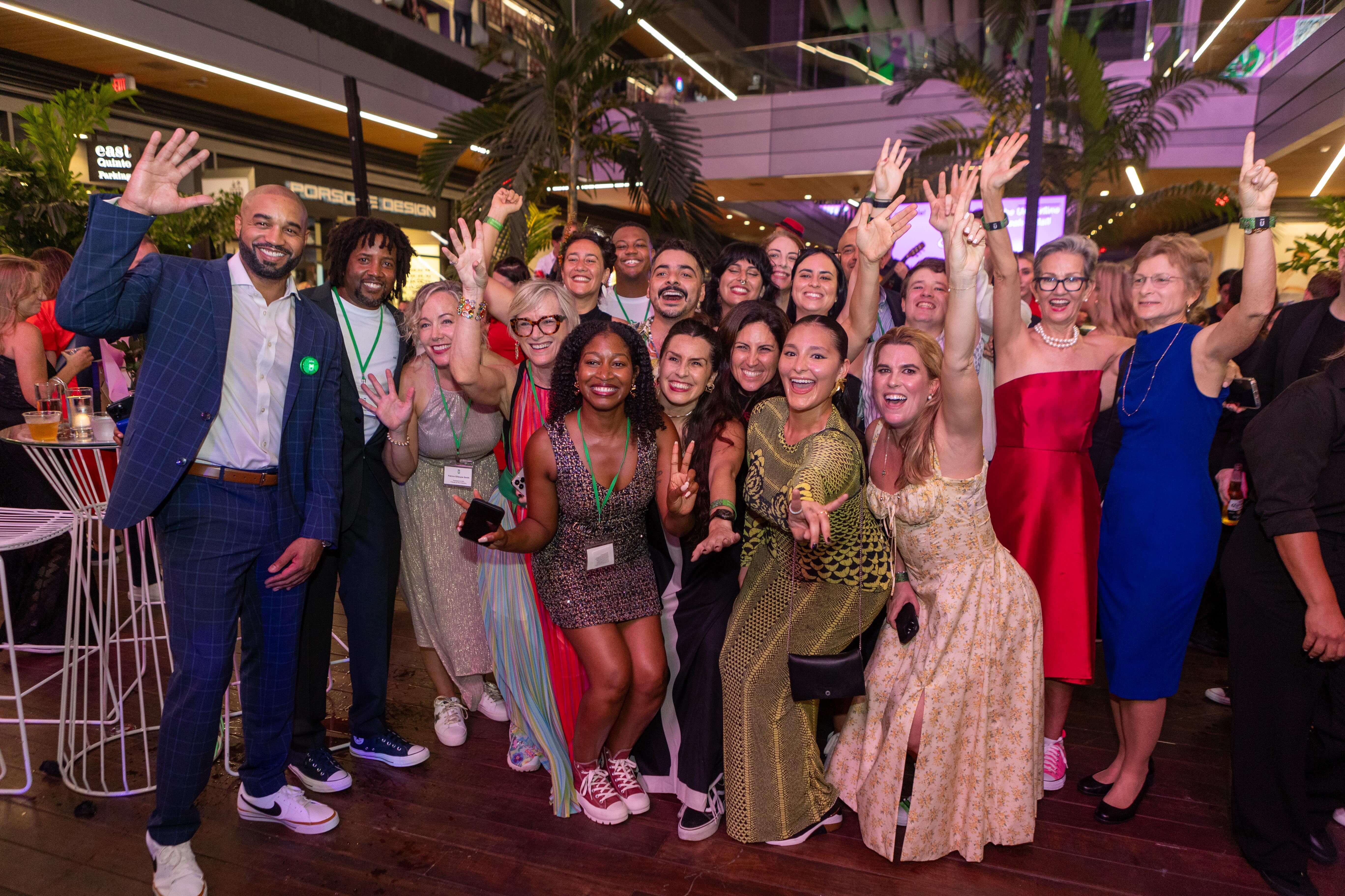 Group of diverse people smiling, posing enthusiastically indoors with raised hands, some wearing event badges.