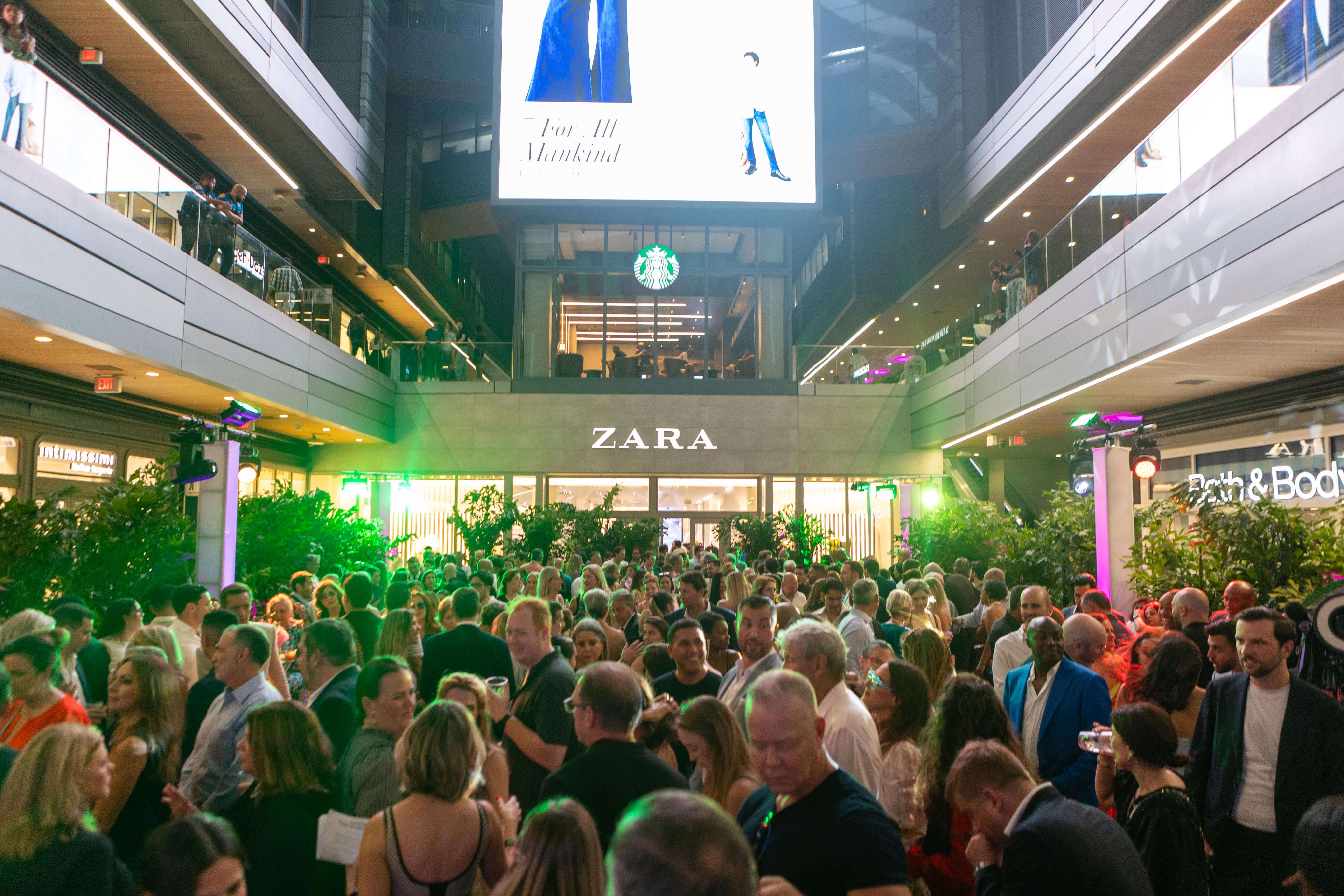 Crowded indoor shopping area with people mingling in front of a Zara store under a Starbucks logo and a large digital screen.