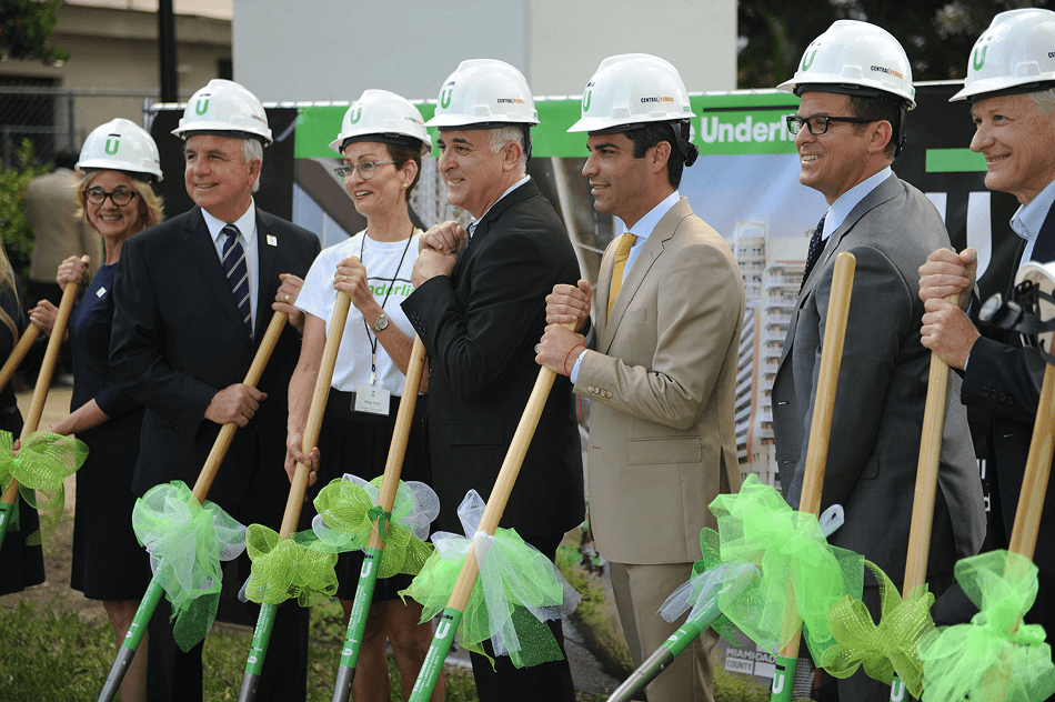 Group of people in hard hats holding shovels decorated with green bows at a groundbreaking ceremony.
