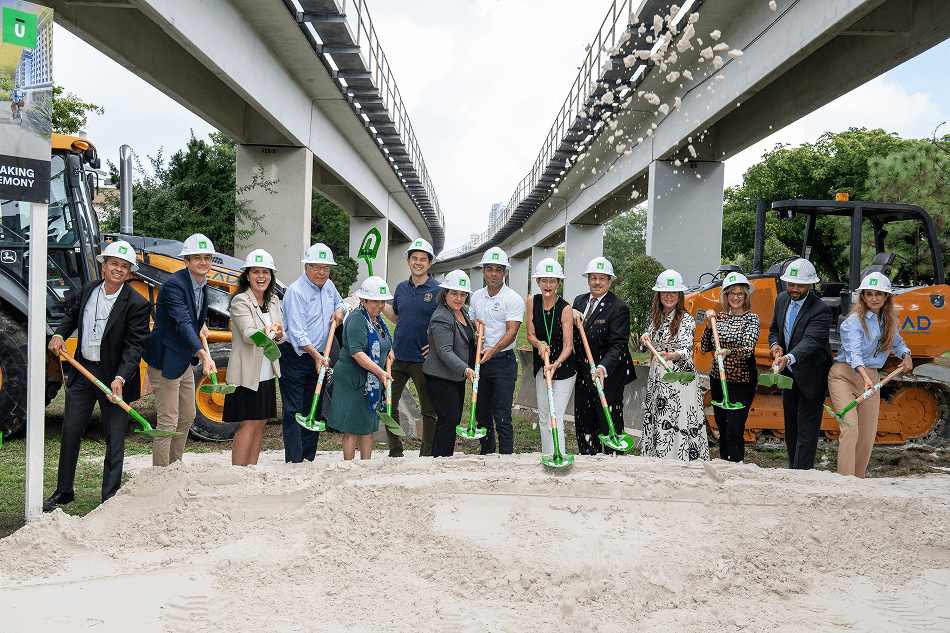 Group of people wearing white hard hats and holding green shovels participate in a groundbreaking ceremony under an elevated railway track with construction equipment in the background.