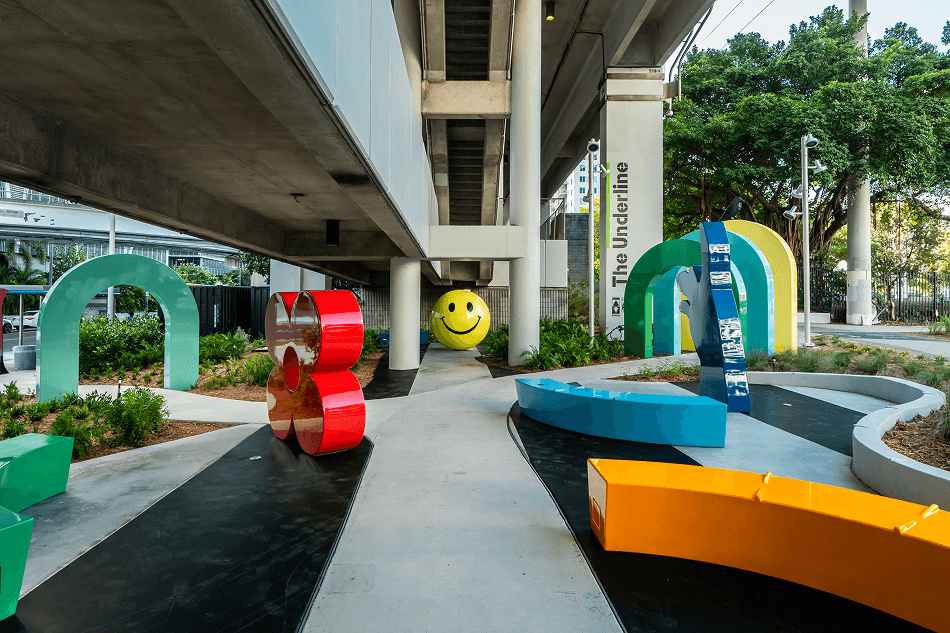 Colorful outdoor playground under a concrete bridge with various curved and numbered structures and a large yellow smiley face ball.