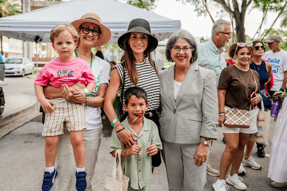 A group of smiling people, including two women holding young children, posing outdoors under a tent at a community event.