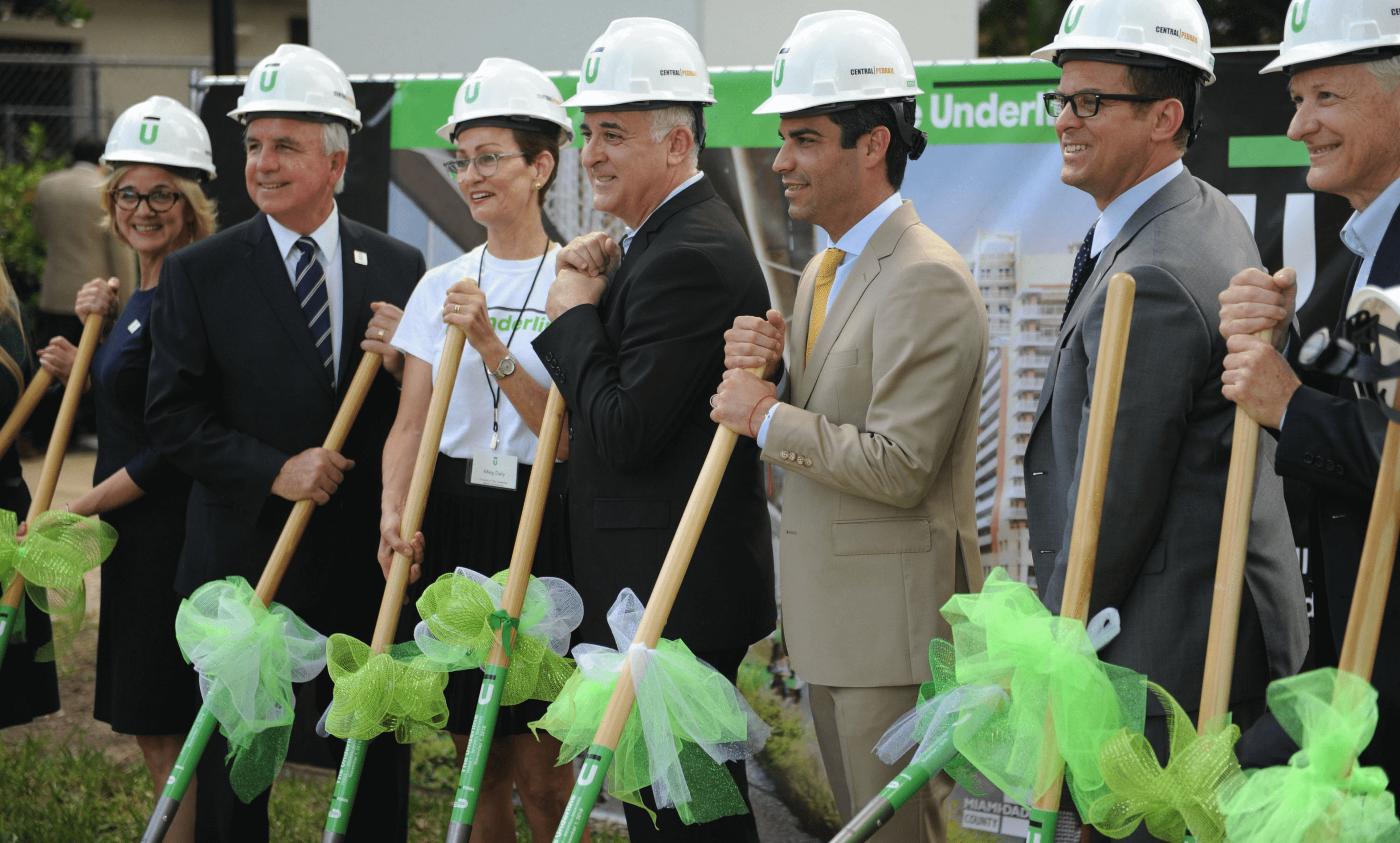 Group of professionals in suits and hard hats holding shovels decorated with green bows at a groundbreaking ceremony.