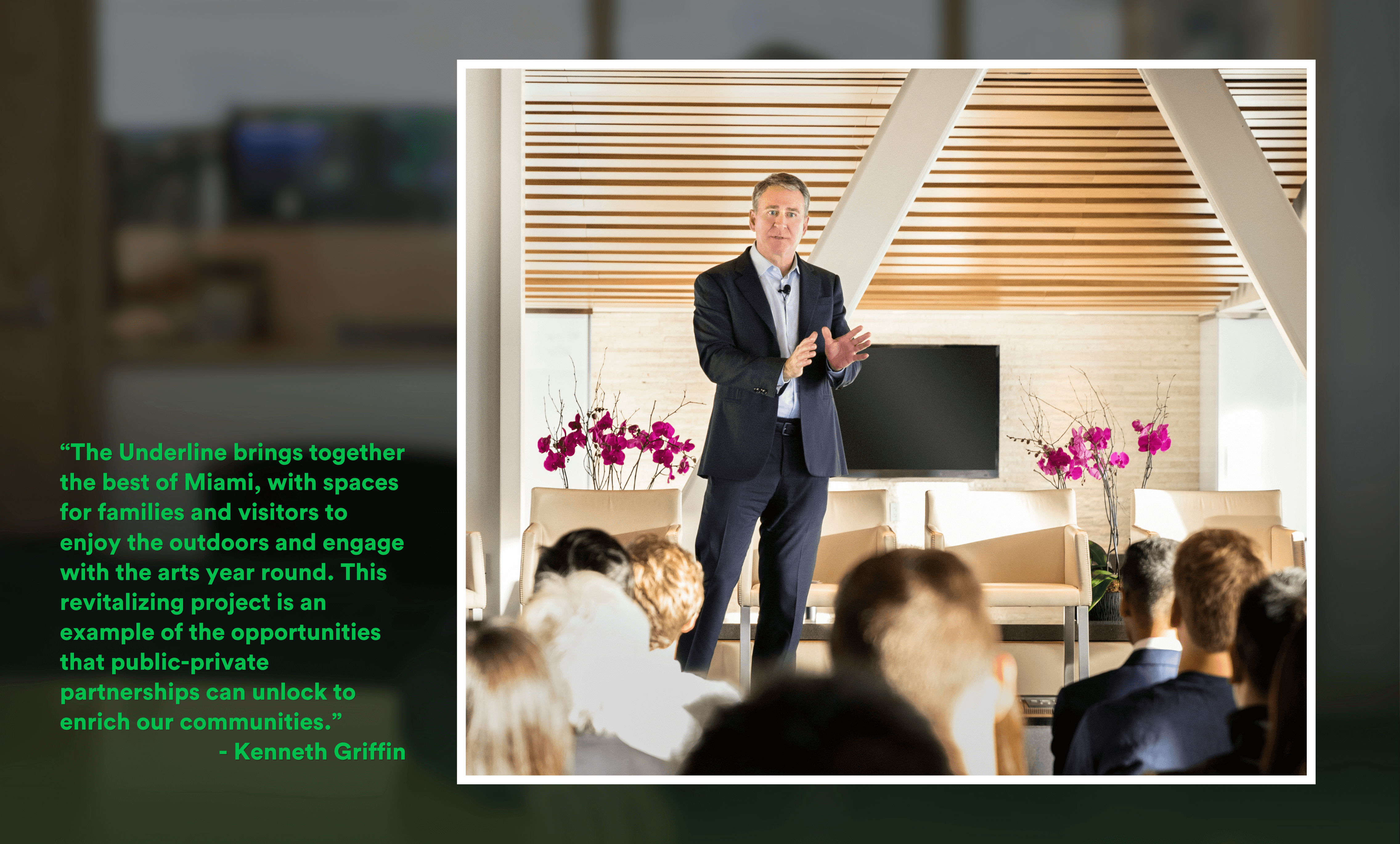 Man in a dark suit speaking to an audience in a modern room with beige chairs and purple orchids on either side.
