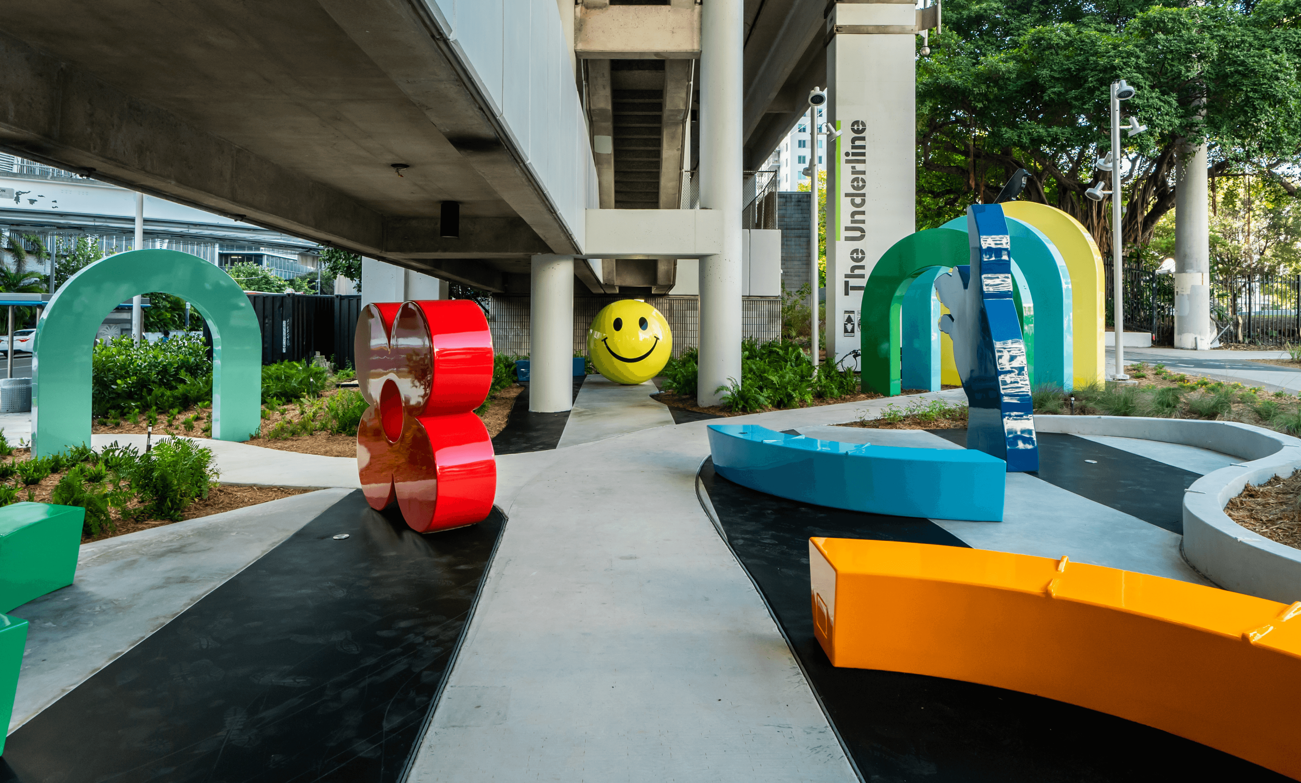 Outdoor area with colorful abstract sculptures, including a large red ampersand, a yellow smiley face, and various blue, green, and orange shapes under a concrete overpass.