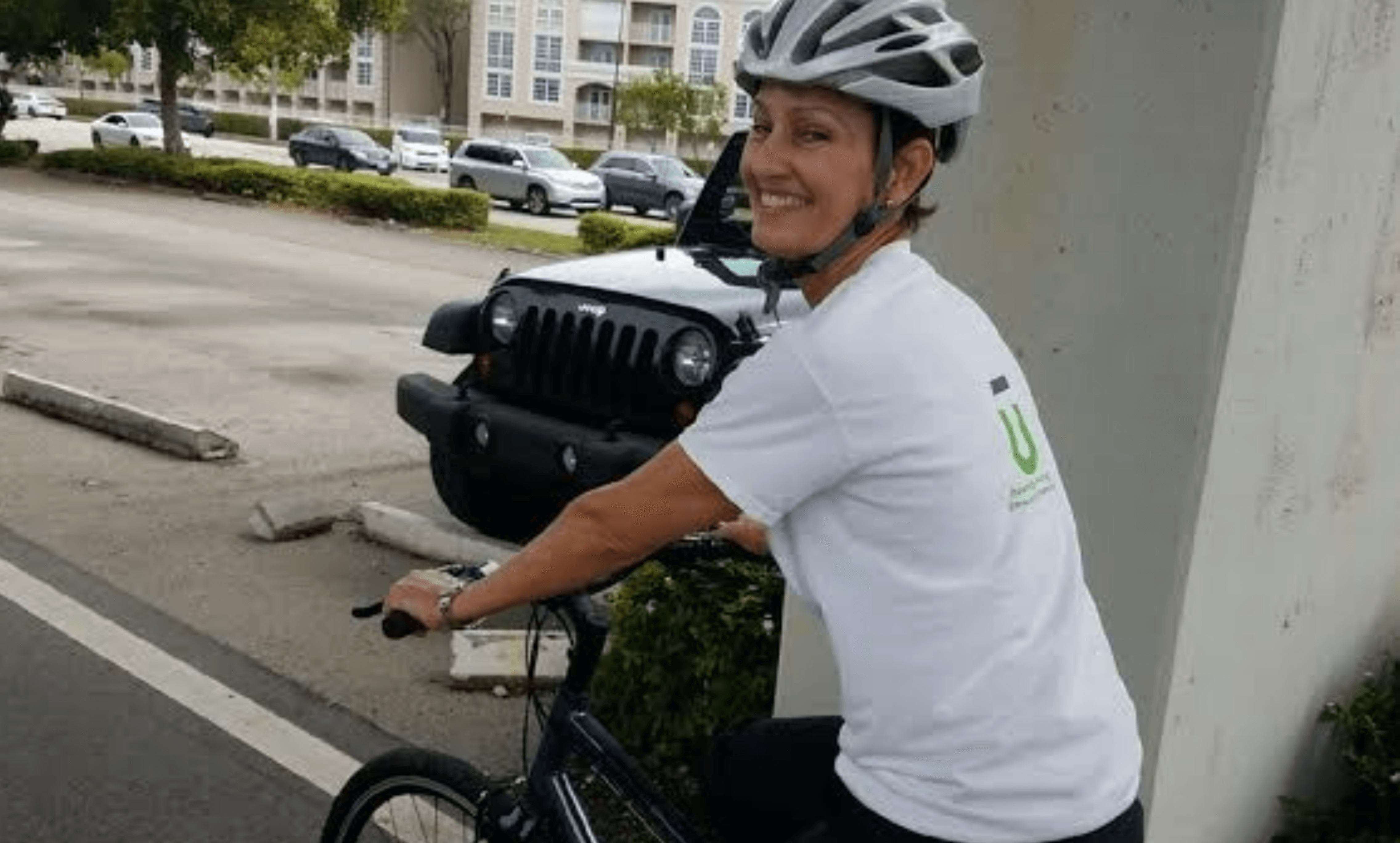 Smiling woman wearing a white helmet and white t-shirt sitting on a bicycle near a parked black Jeep in an outdoor parking area.