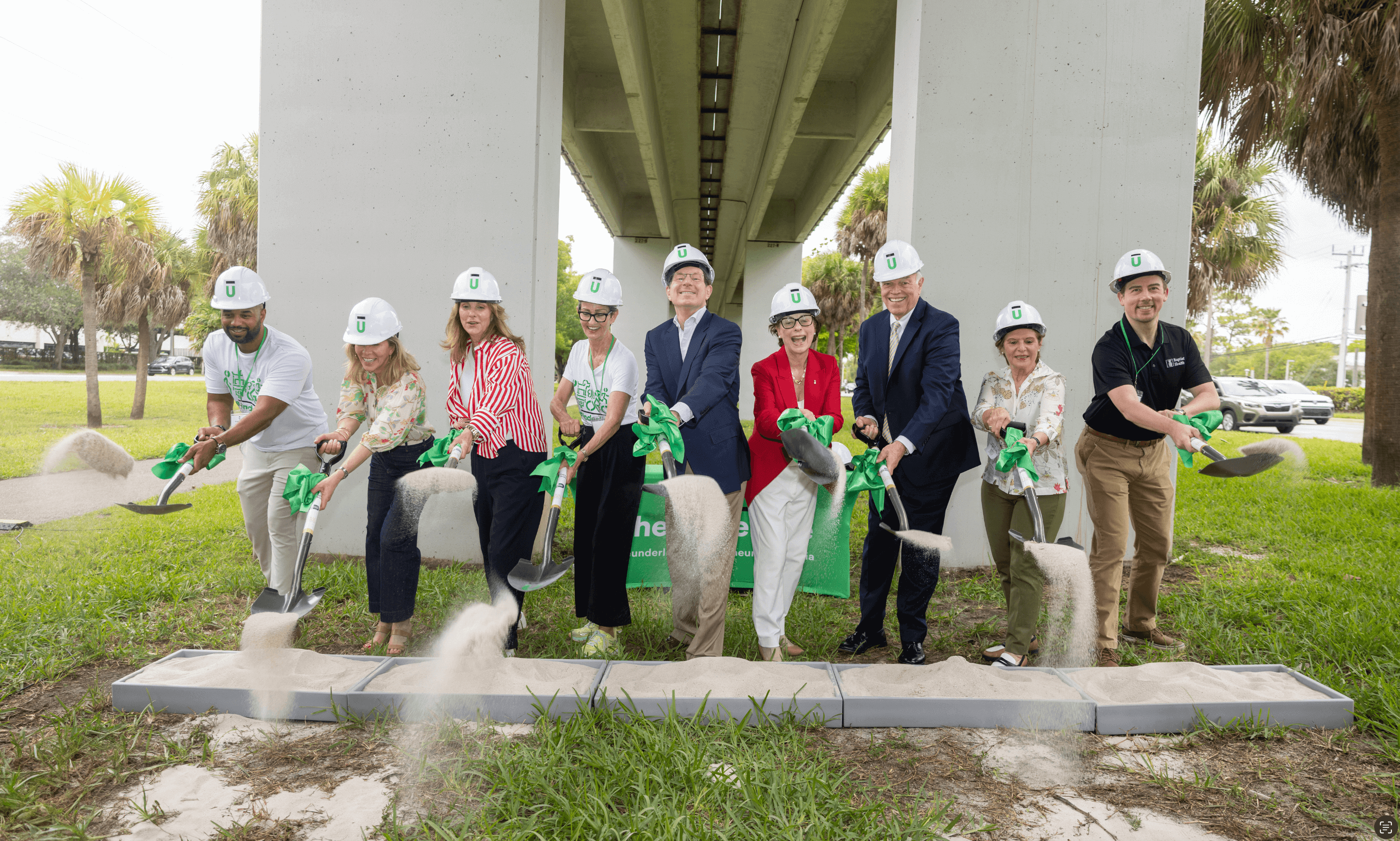Nine people wearing white hard hats with a green U logo are participating in a groundbreaking ceremony under a bridge, shovel sand into a box in front of them.