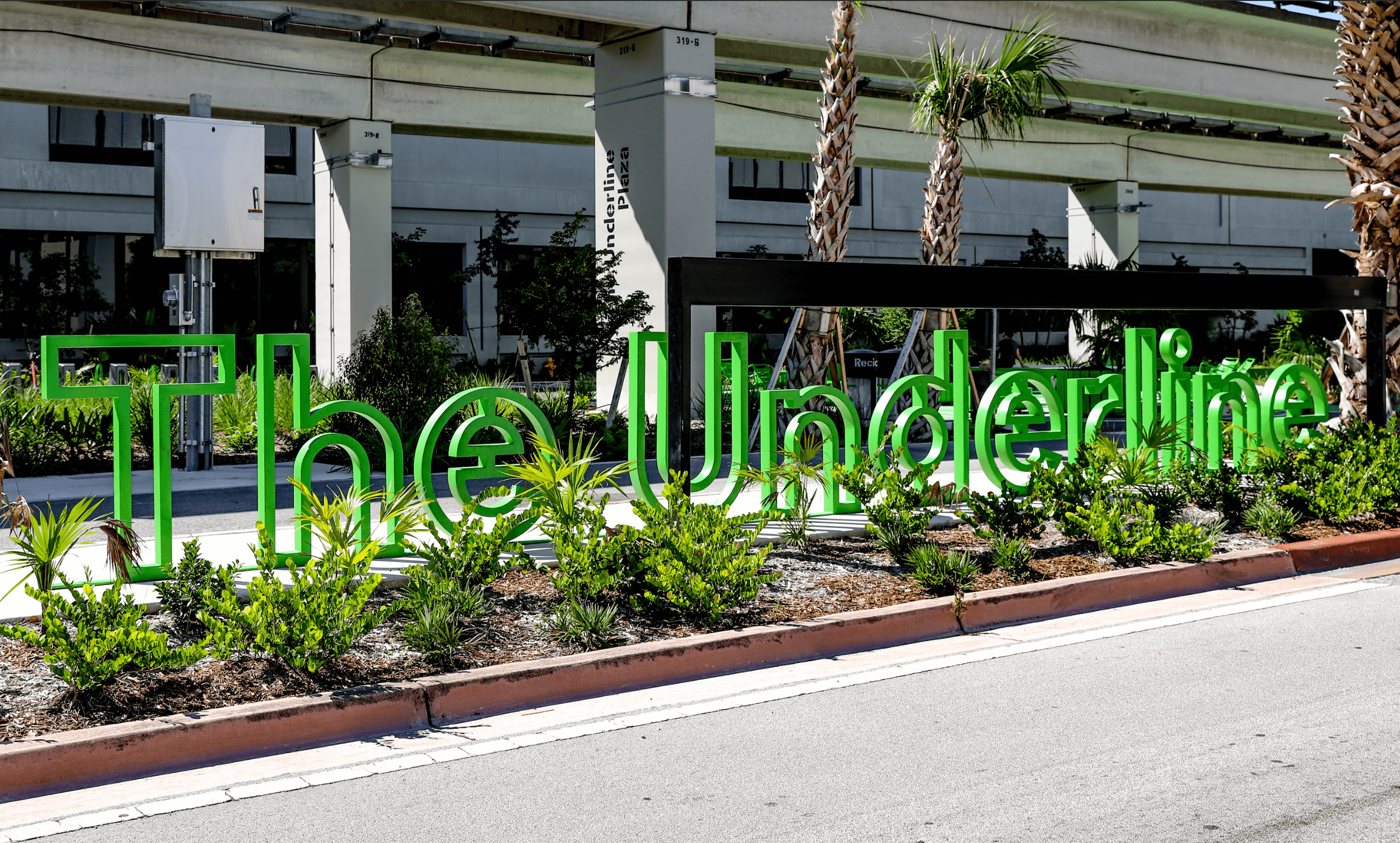 Green open-frame letters spelling 'The Underline' surrounded by plants and palm trees near elevated train tracks.