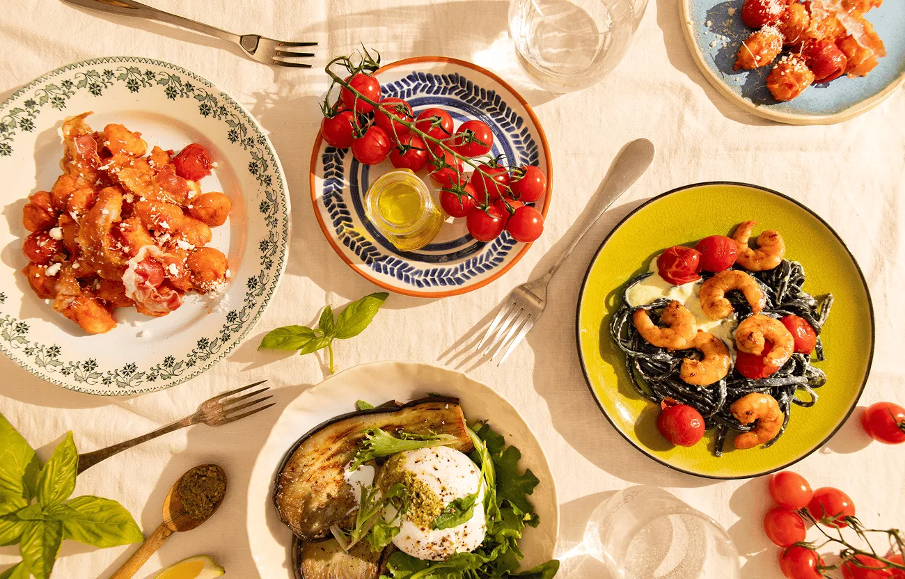Table dressée avec différents plats cuisinés par Popchef : gnocchis à la tomate, pâtes aux crevettes, burrata avec légumes grillés et tomates cerises
