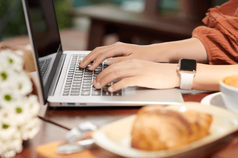 Hands of woman typing on laptop with croissant and coffee
