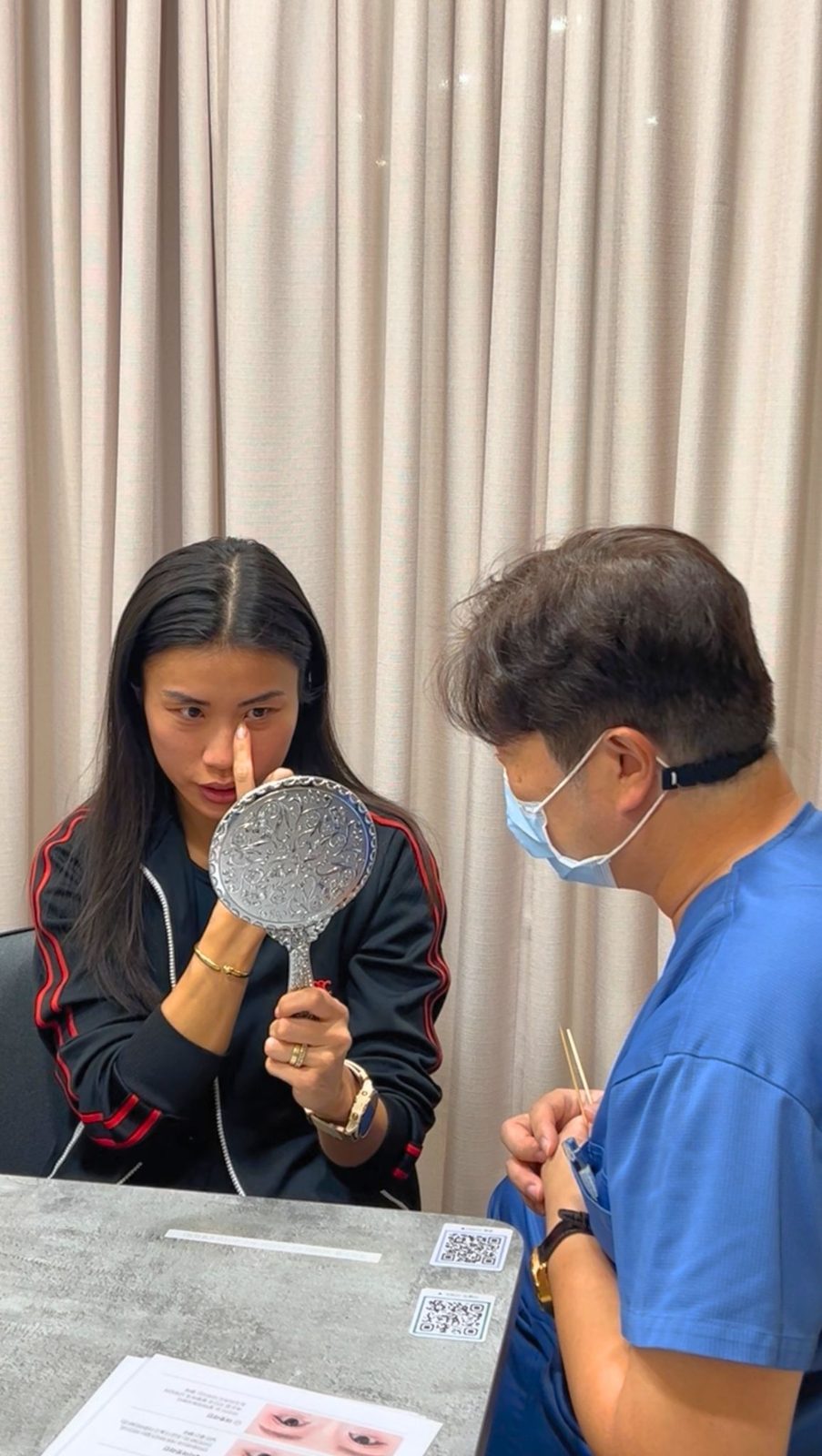 A patient holding a hand mirror while a doctor marks her skin during a consultation