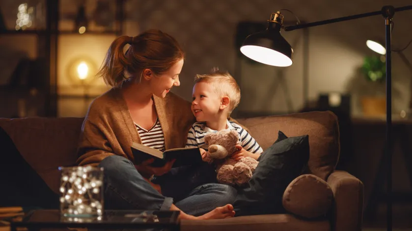 En mamma och ett barn som sitter på soffan och läser i kvällsbelysning.