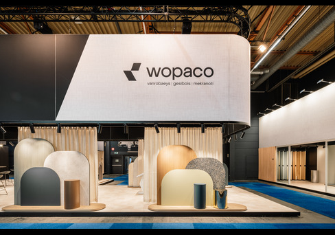 Modern trade show booth for Wopaco with curved panels and neutral tones under black ceiling beams.