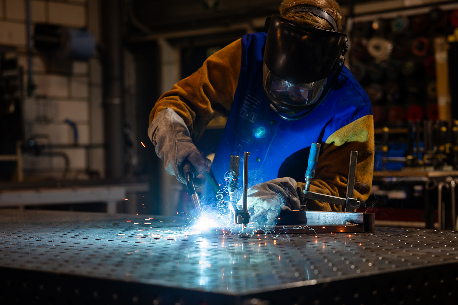 Worker with protective gear welding metal on a large workshop table with sparks flying.