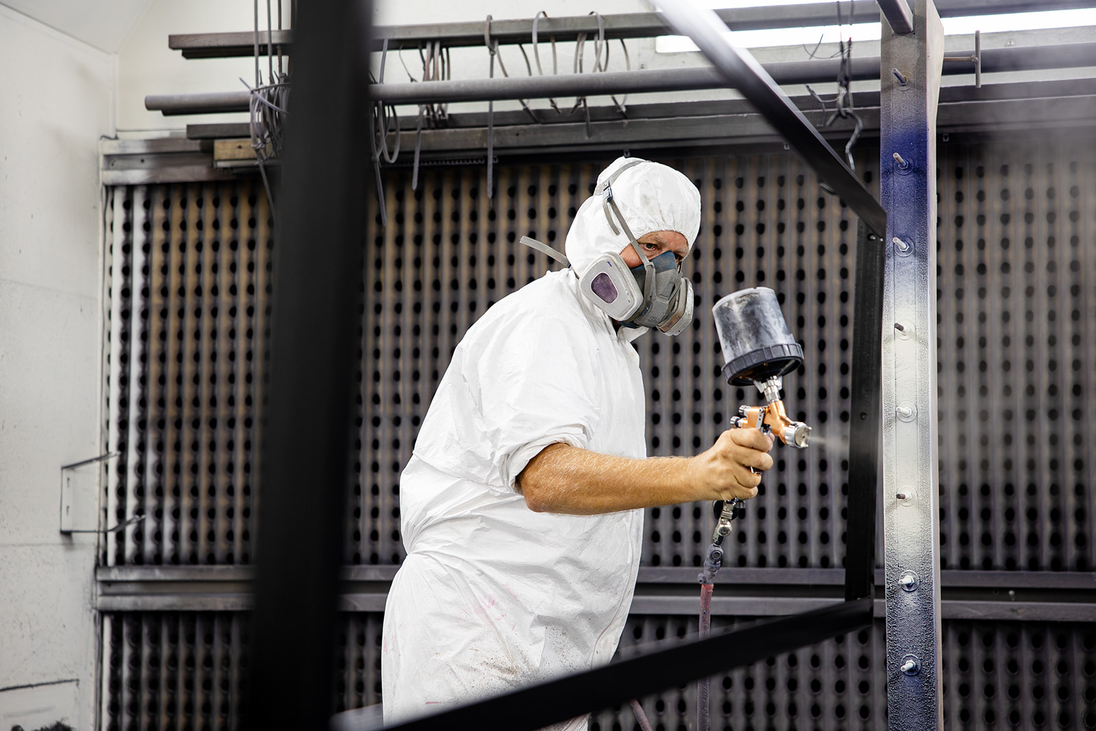 Worker in white protective suit and mask spray-painting a metal frame indoors.