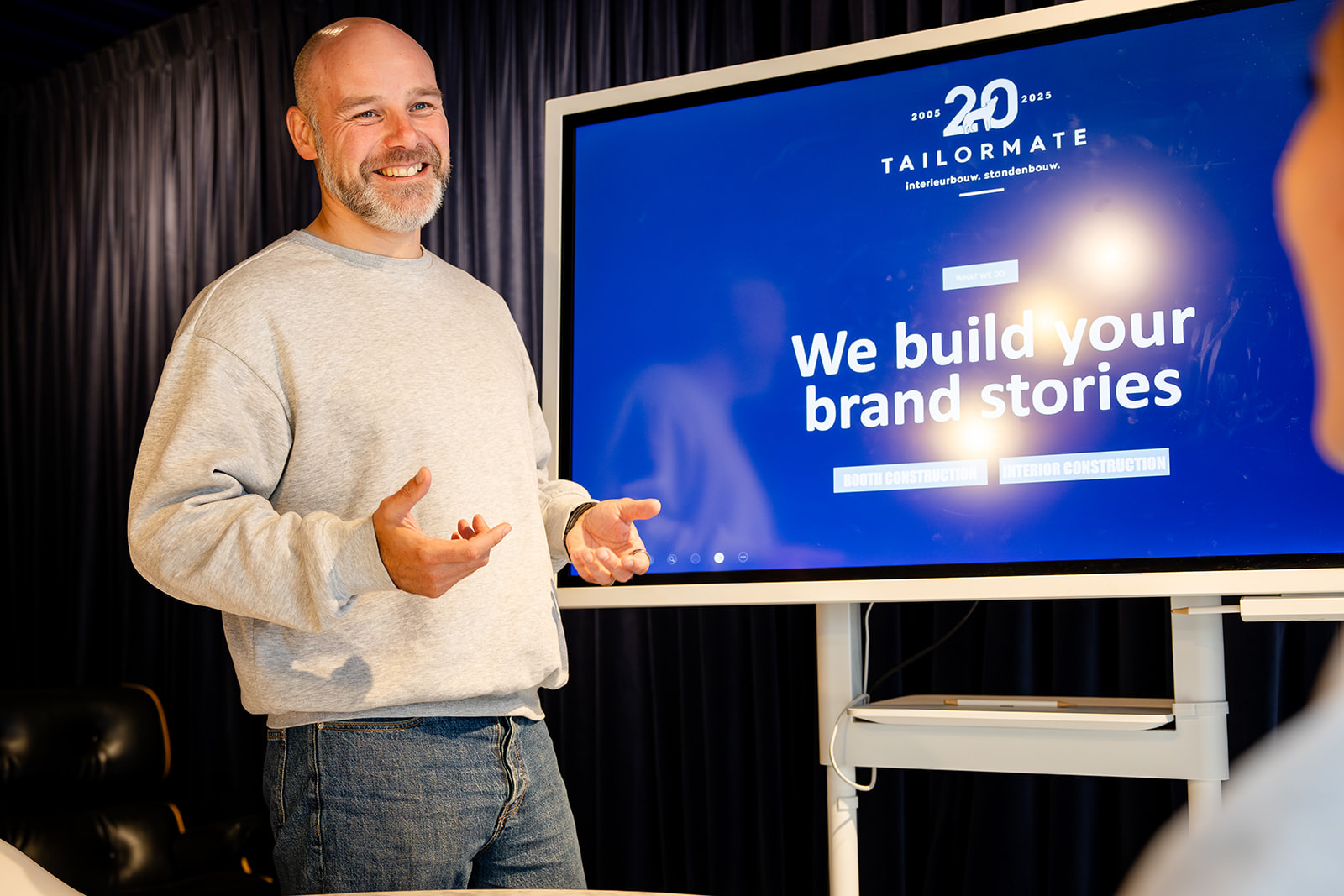 Smiling man in grey sweater presenting in front of a screen showing Tailormate branding message.
