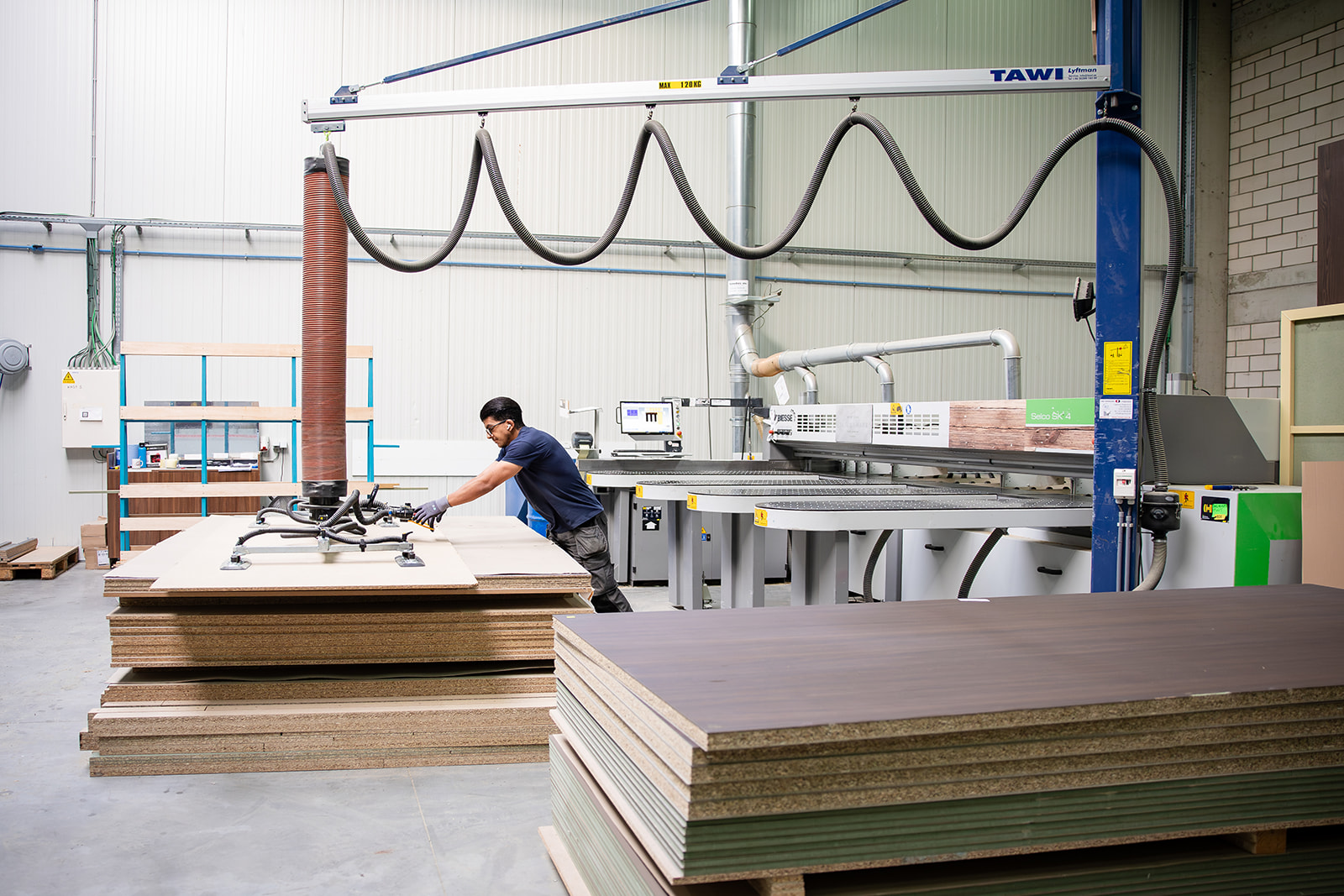 Worker handling large wood panels with industrial vacuum lifter in a factory setting.