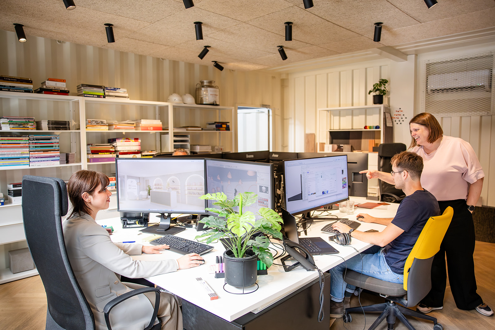 Two people working at desks with multiple monitors in a bright modern office with shelves and plants.