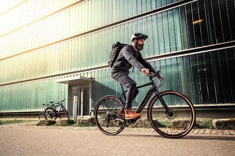 Man wearing helmet and backpack riding a black bicycle on a street beside a large glass building.