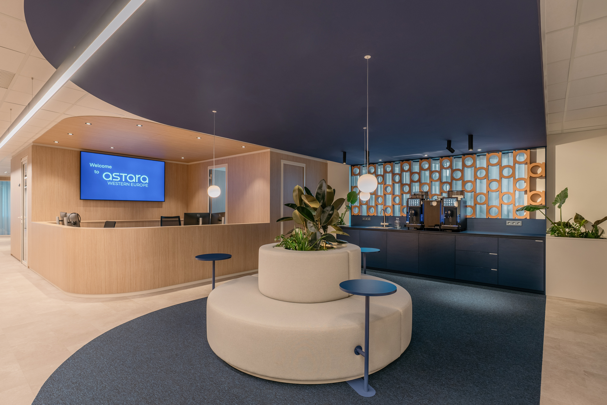 Modern office reception area with curved desk, digital welcome sign, central seating with plants, and coffee machines.