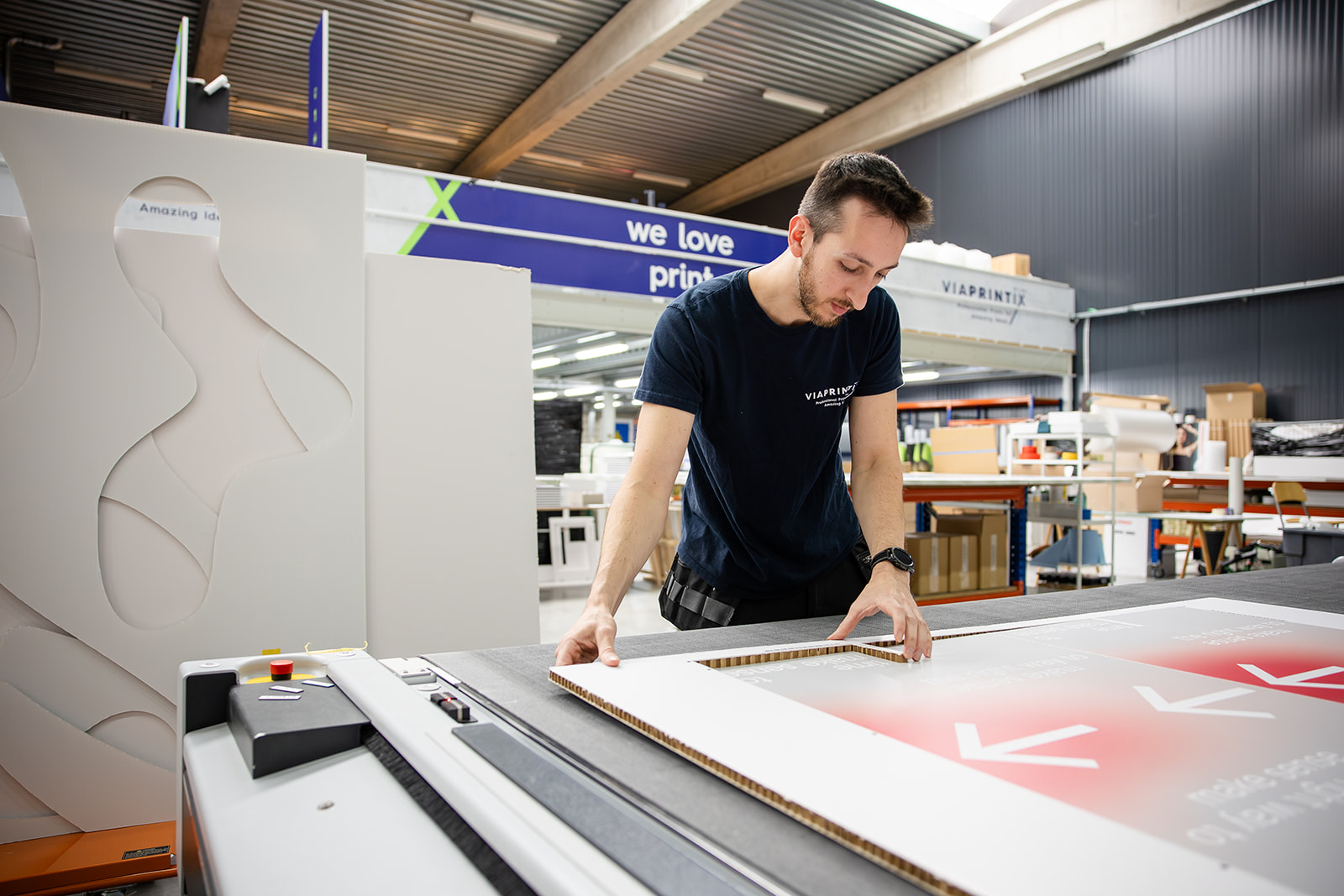 Man inspecting a large printed sheet with cutouts in a busy print shop warehouse.