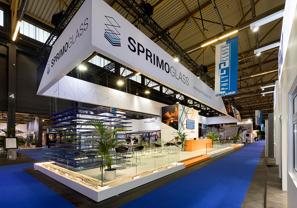 Sprimoglass exhibition stand with glass panels, seats and blue floor in a spacious hall.