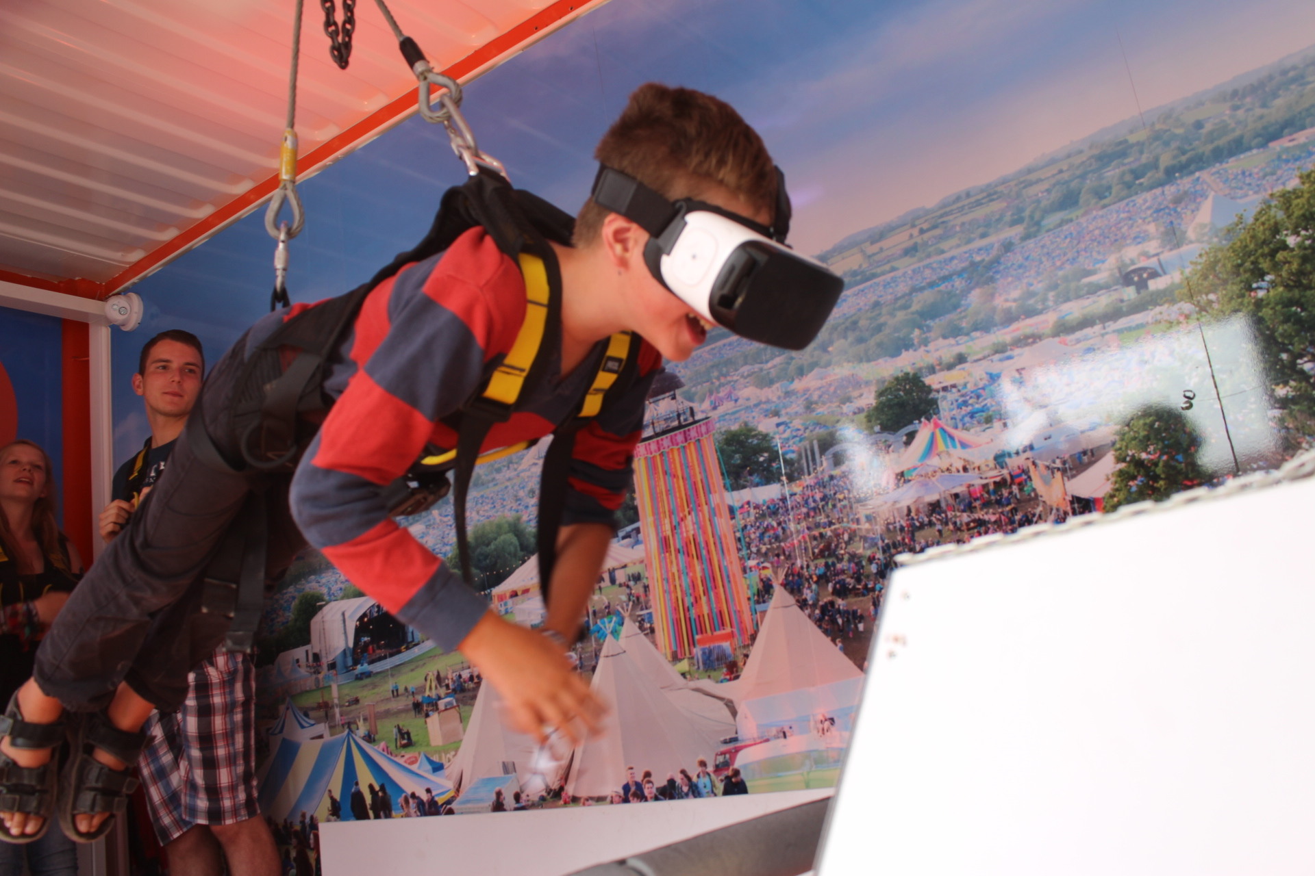 Boy with VR headset harnessed and suspended simulating flying over a festival backdrop.