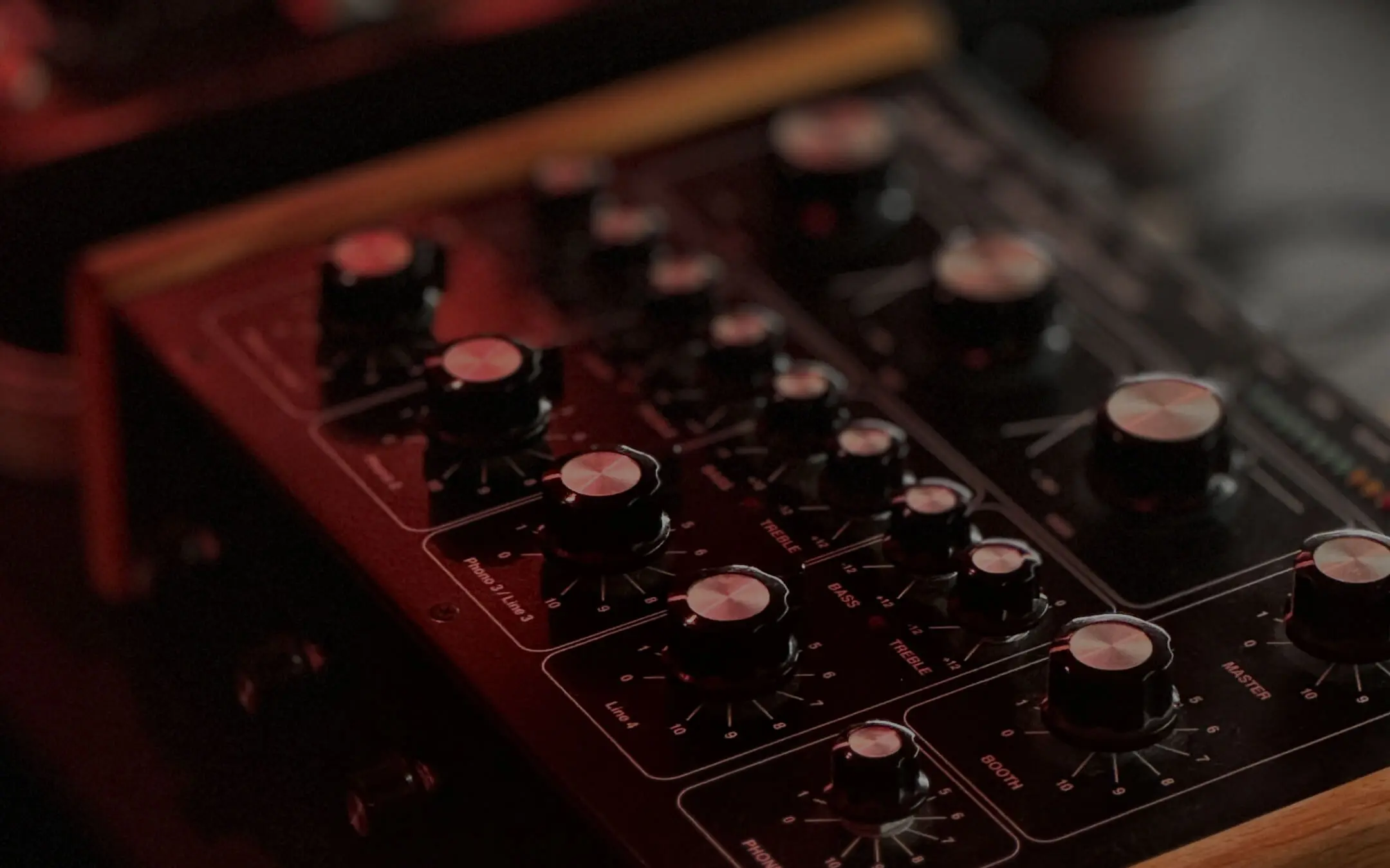 Close-up of a DJ mixer with multiple control knobs for volume, bass, treble, and other audio settings under low red lighting.