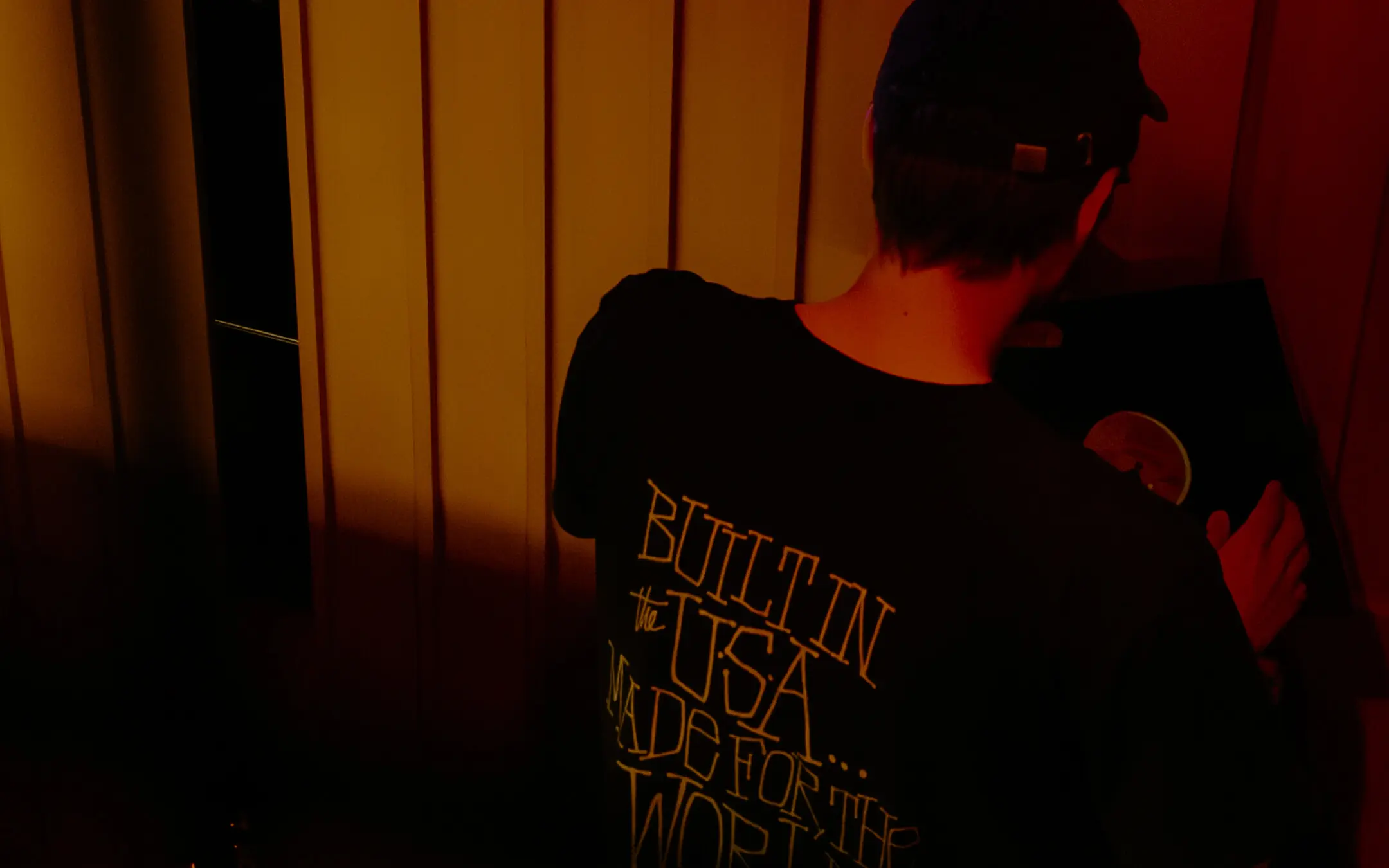 Person wearing a black cap and black shirt holding a vinyl record, shirt text reads 'BUILT IN the USA MADE FOR THE WORLD.'