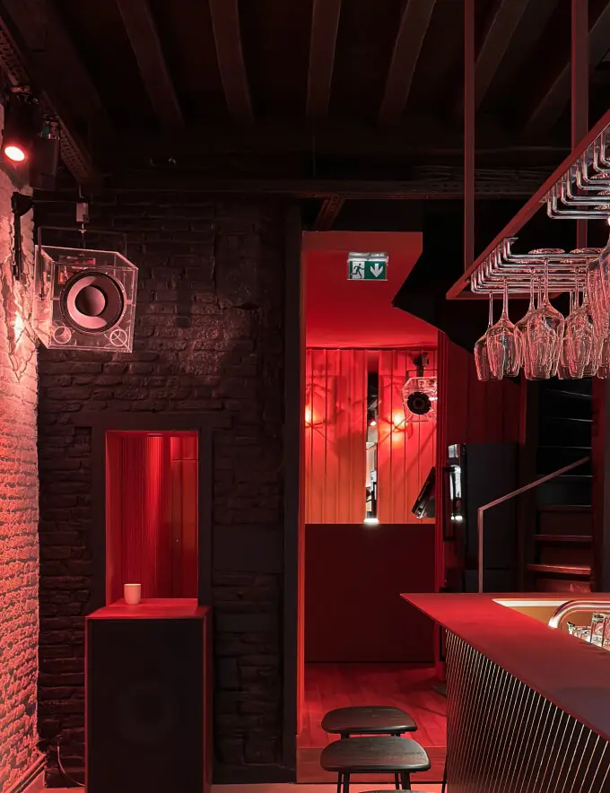 Dimly lit bar area with red lighting, brick walls, hanging wine glasses, and two stools near the counter.