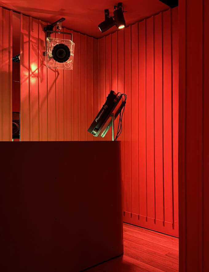 Dimly lit DJ booth with a Pioneer CDJ player, speaker mounted on a wall, and red vertical paneling under red lighting.