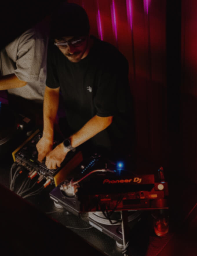 DJ wearing a black shirt and beanie adjusting audio mixer knobs in a dimly lit room with red and purple lights.