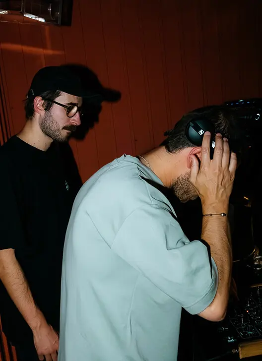 Two men, one wearing glasses and a black cap, the other adjusting headphones near DJ equipment against a wooden wall backdrop.
